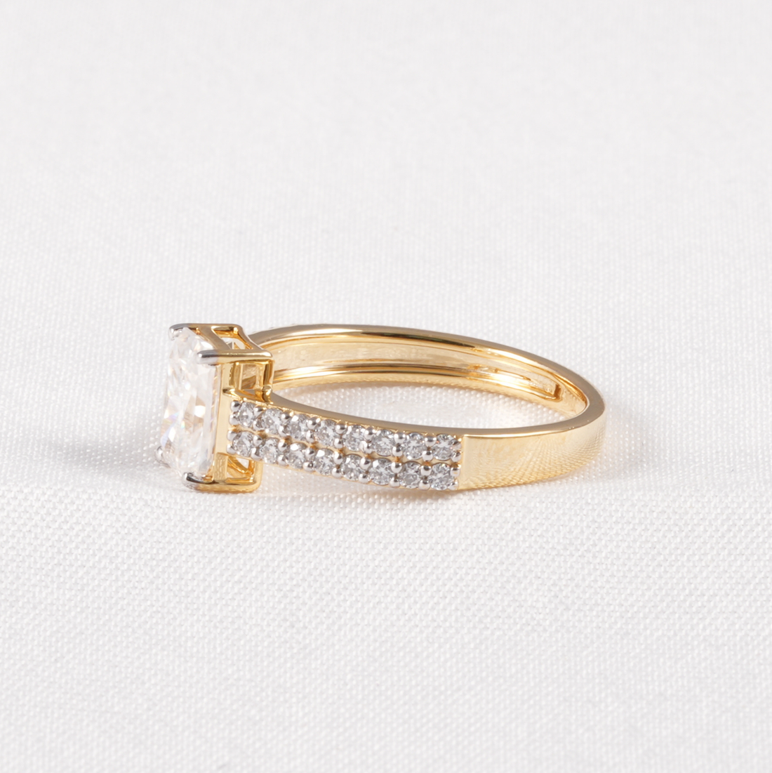 A yellow gold ring featuring a large rectangular clear gemstone in a prong setting, with a pavé band accented by small clear stones, photographed on a textured white fabric background.