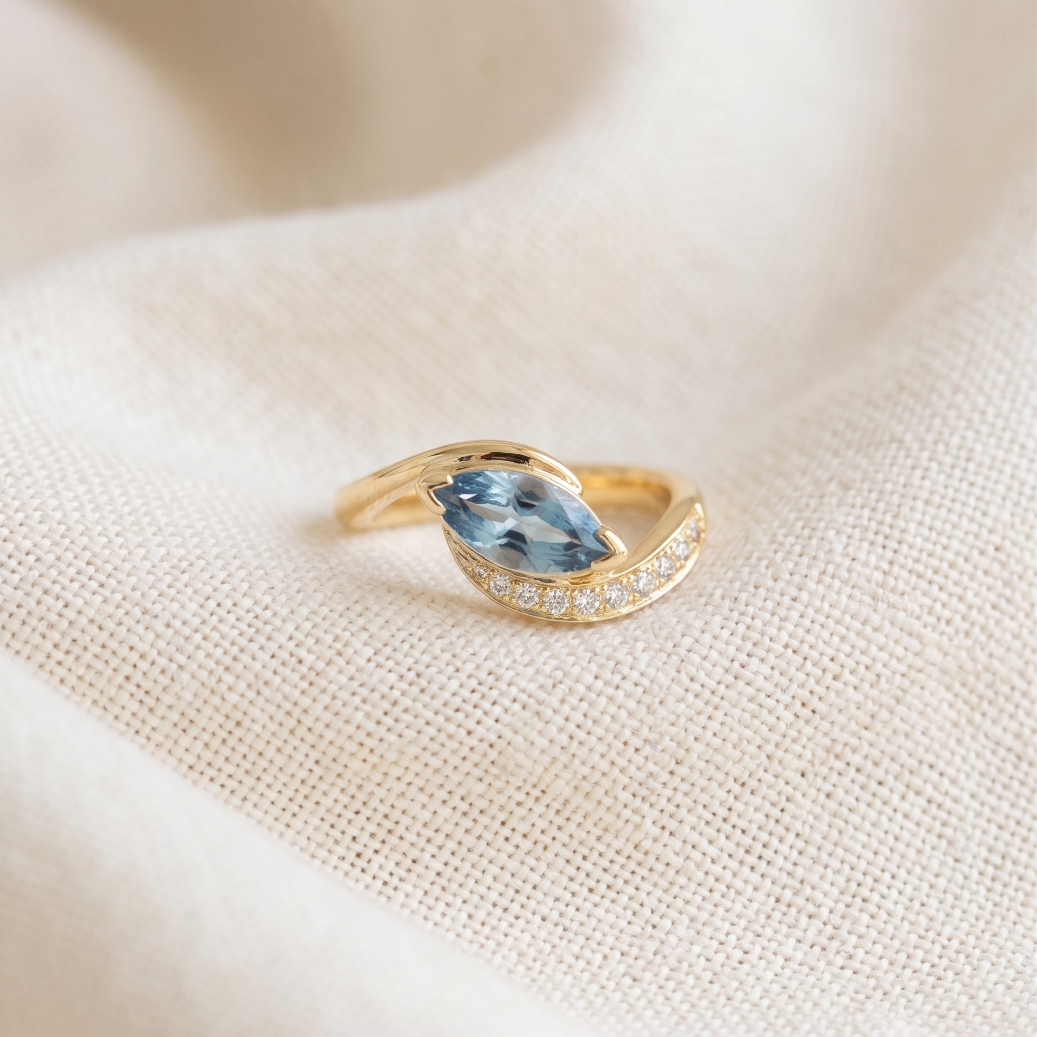 A gold ring featuring a marquise-cut light blue gemstone and a curved row of small clear stones, resting on a textured beige fabric background.