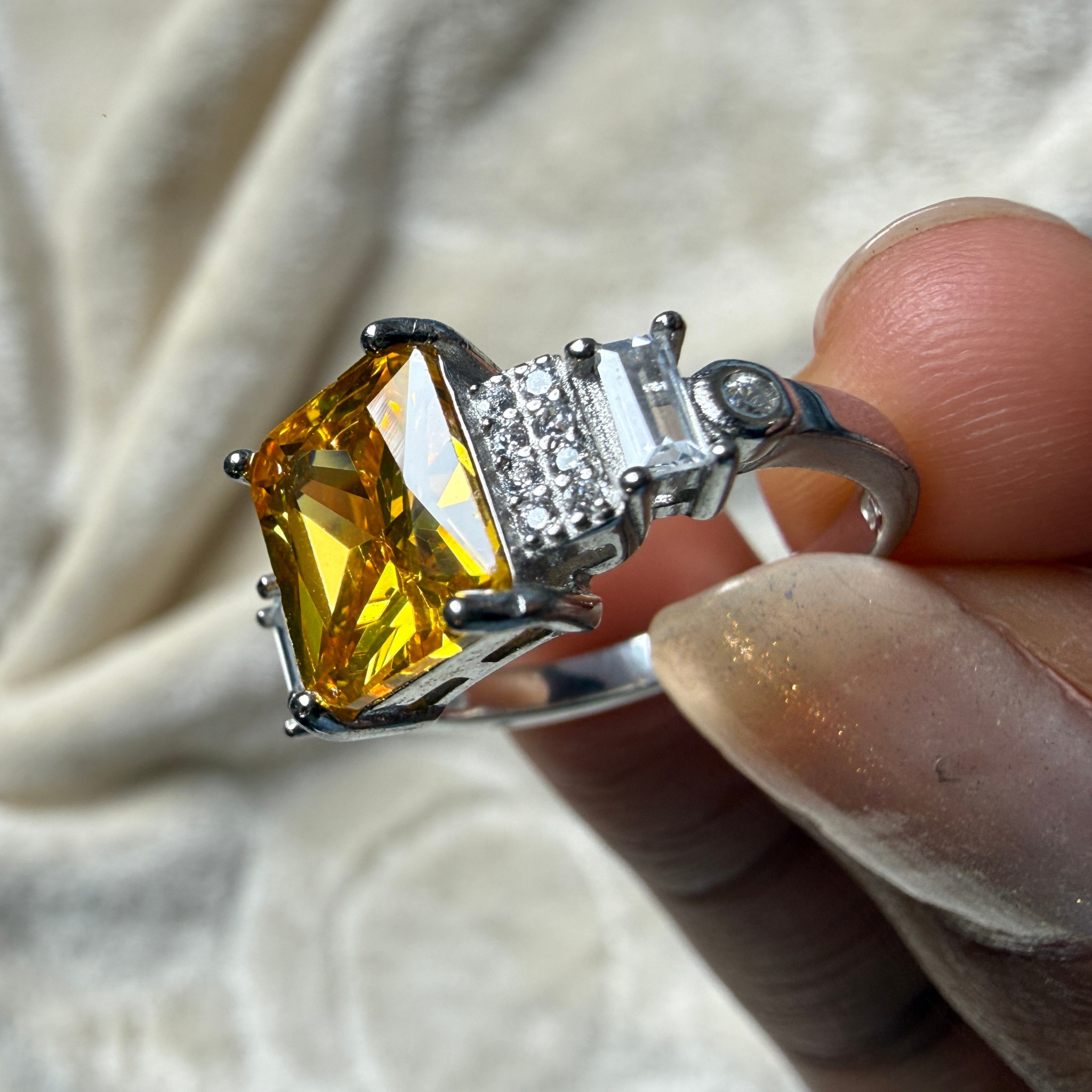 A silver ring featuring a large, faceted yellow gemstone and a side section with small clear stones, held by a hand against a soft, textured cream fabric background.