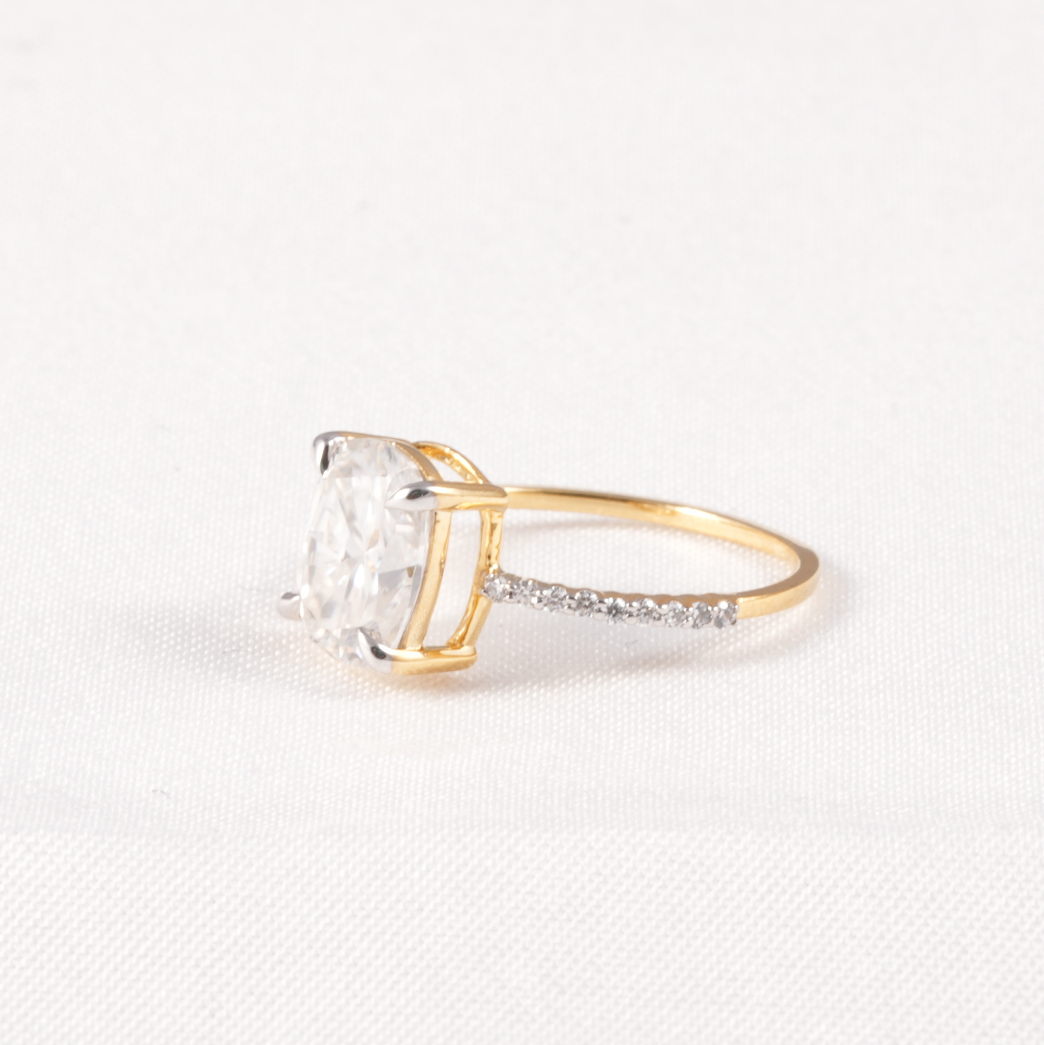 A yellow gold ring featuring a large cushion-cut clear gemstone in a prong setting, with a pavé band accented by small clear stones, shown on a textured white background.