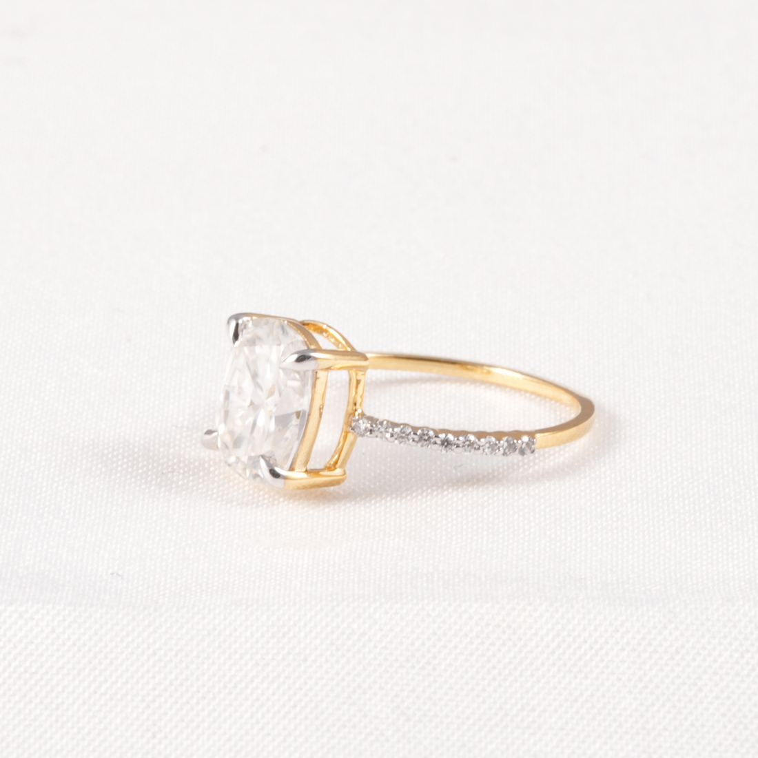 A yellow gold ring featuring a large cushion-cut clear gemstone in a prong setting, with a pavé band accented by small clear stones, shown on a textured white background.