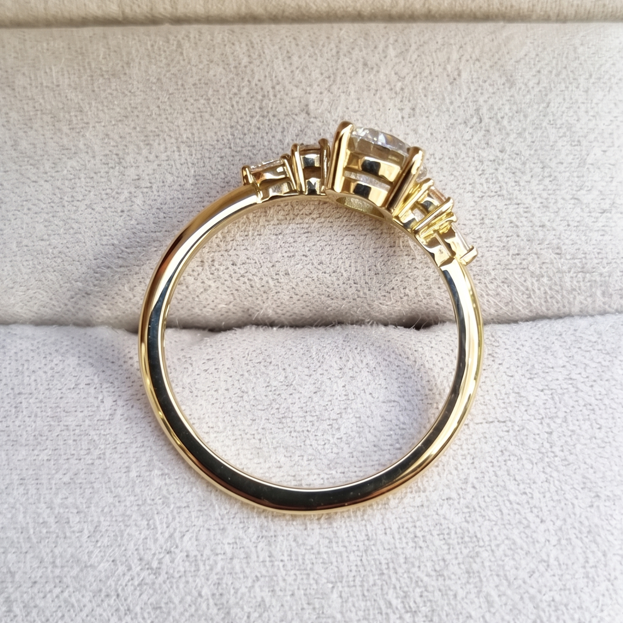 A yellow gold ring with a round-cut center stone and smaller side stones, displayed on a textured light beige fabric surface.