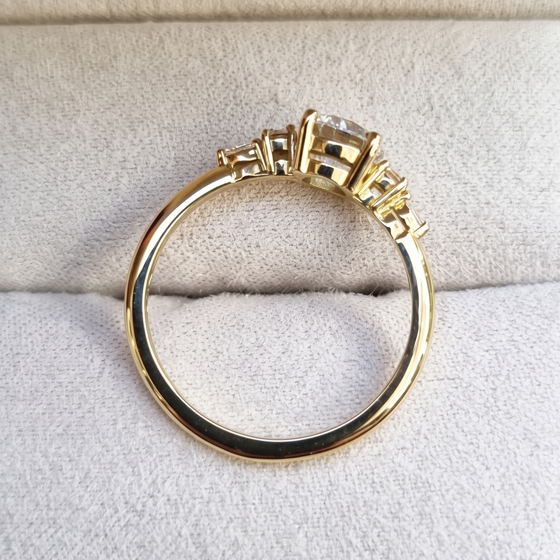 A yellow gold ring with a round-cut center stone and smaller side stones, displayed on a textured light beige fabric surface.
