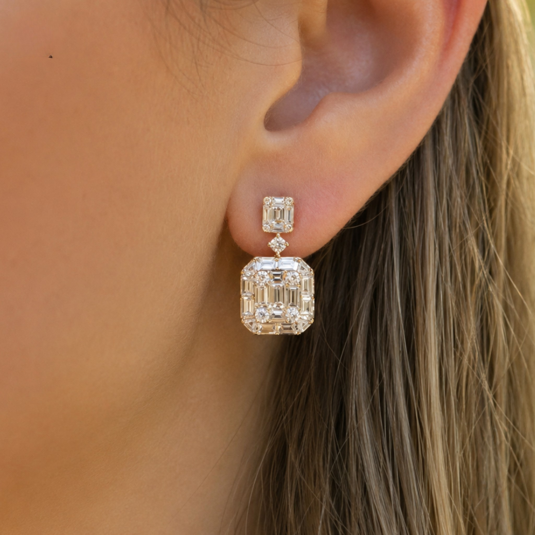 A close-up of a model's ear wearing a drop earring with a large emerald-cut clear gemstone in a silver-toned setting.
