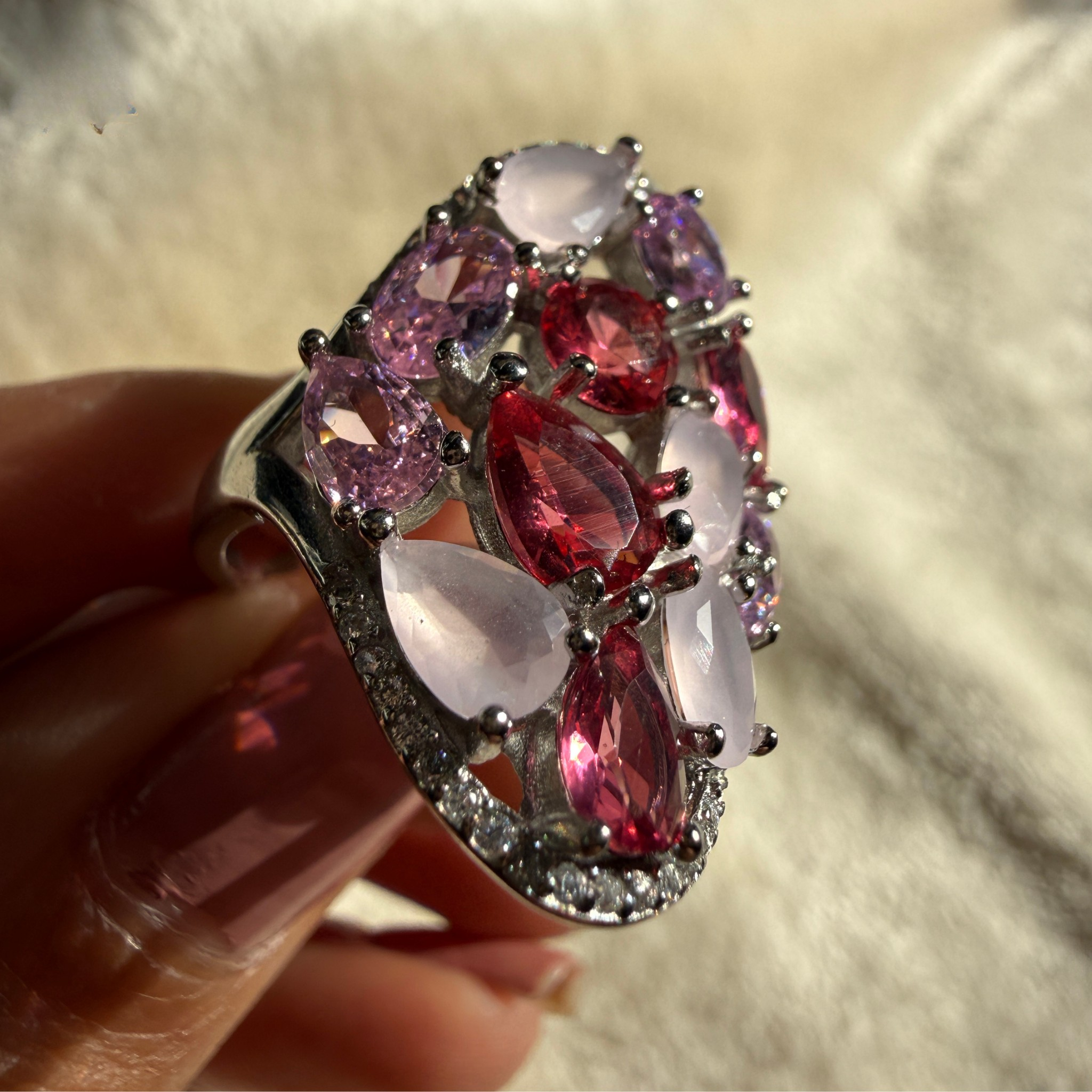 A close-up of a hand holding a large statement ring with marquise and oval pink gemstones in a silver-toned setting, against a soft beige fabric background.