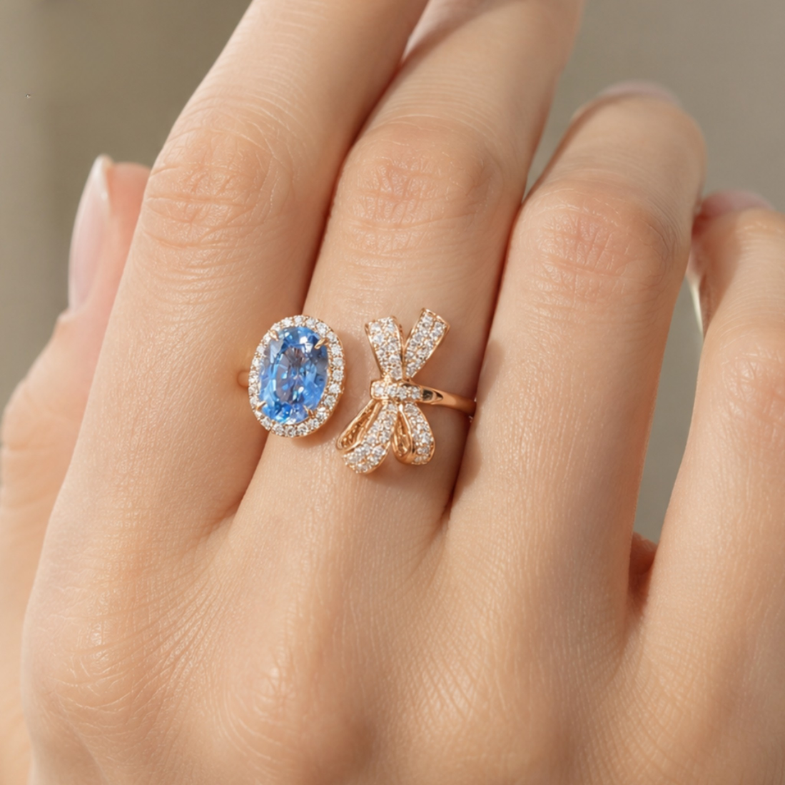 A close-up of a hand wearing two rose gold rings: one with an oval-cut blue sapphire surrounded by small clear stones, and the other shaped like a bow with pavé-set clear stones.