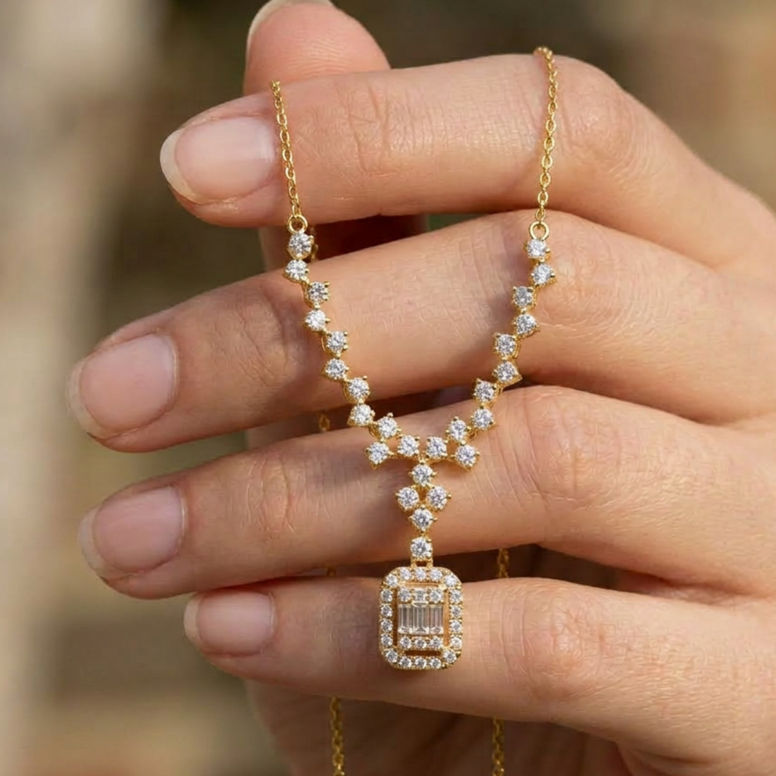 A close-up of a hand holding a delicate gold necklace with a rectangular emerald-cut pendant surrounded by small clear stones.