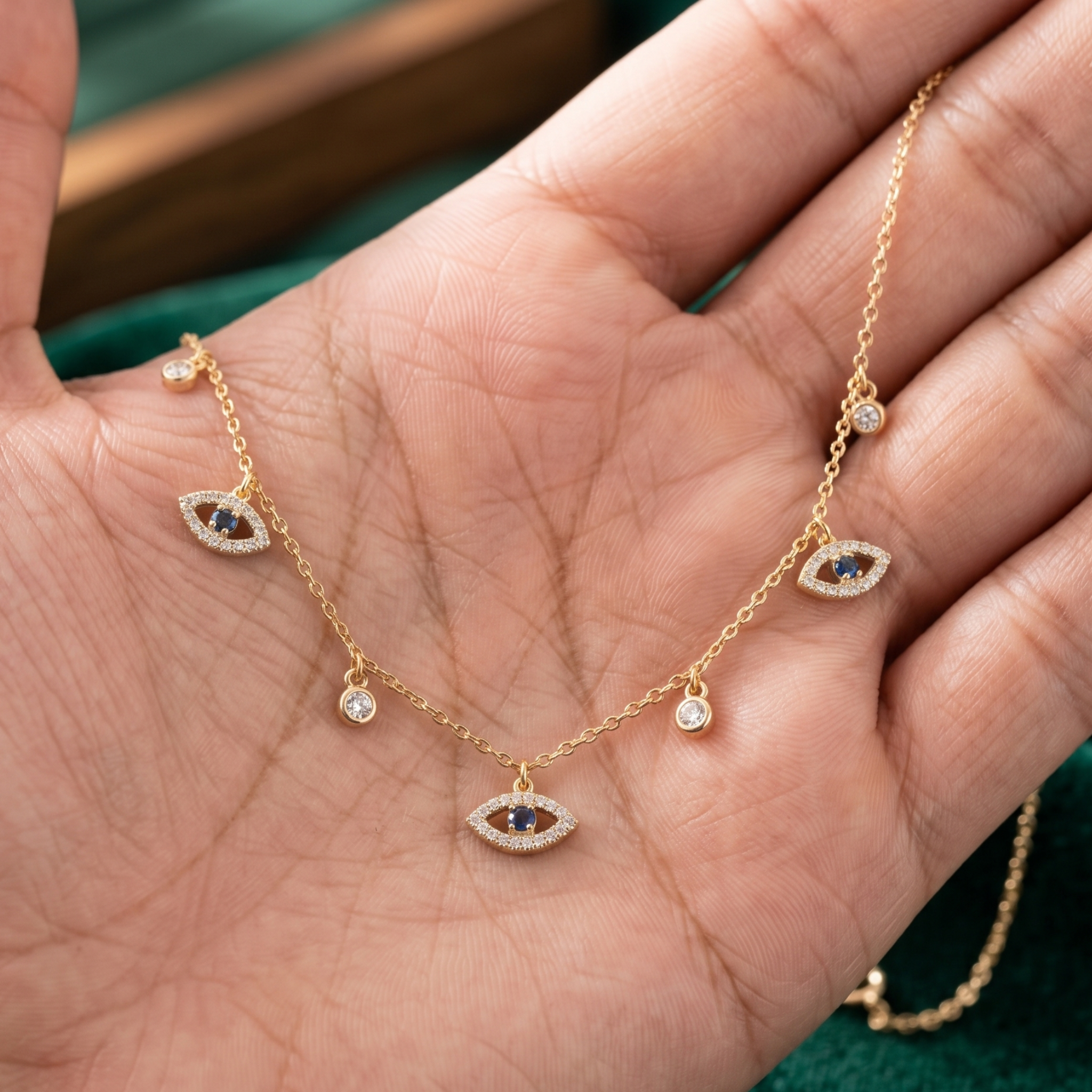 A gold chain necklace with five evil eye charms, held in a person's hand. The evil eye charms feature dark blue gemstone centers surrounded by small clear stones, and the necklace is shown against a background of skin and a dark green surface.