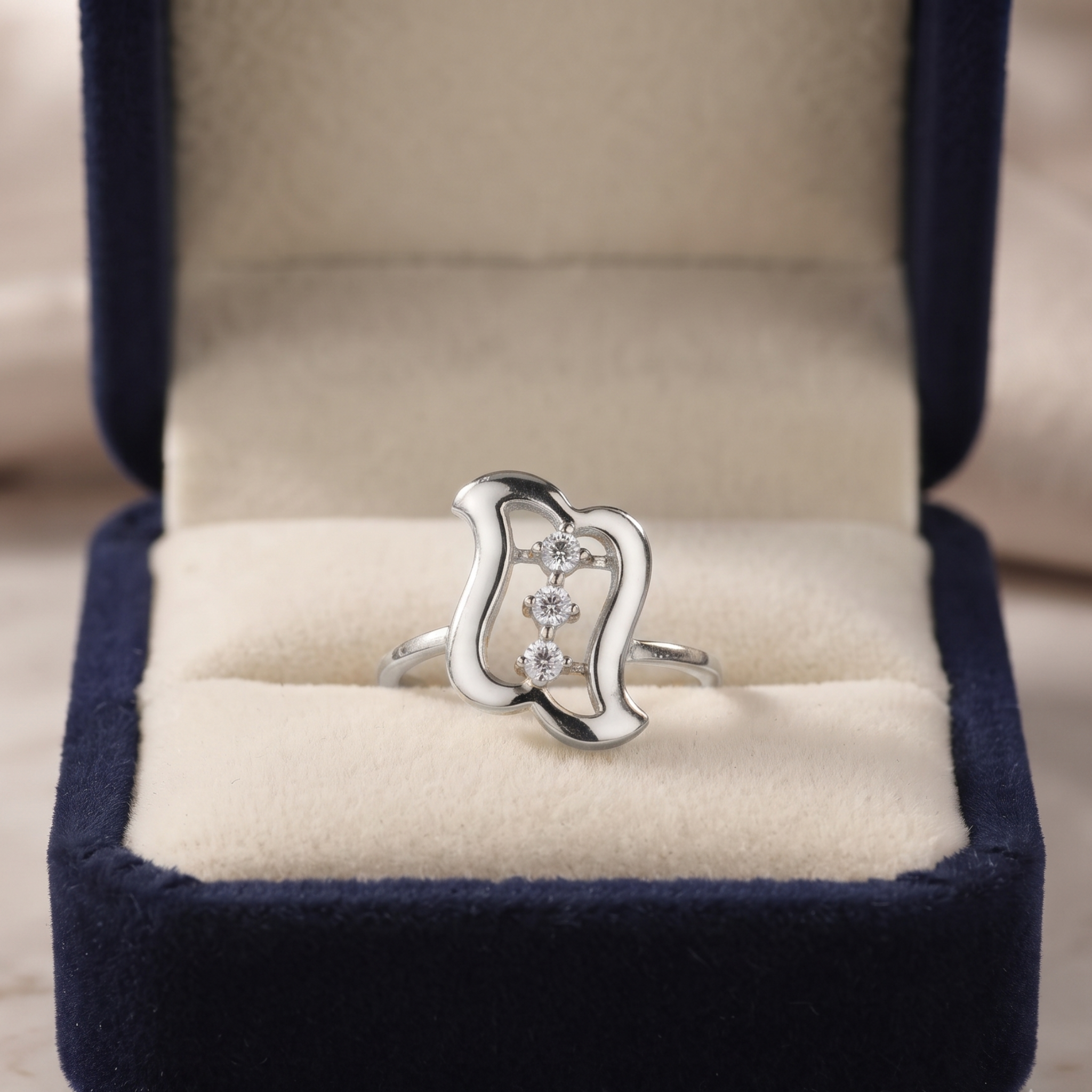 A silver ring with three small clear stones set in an abstract open-loop design, displayed inside an open navy blue velvet jewelry box with a cream-colored cushion.