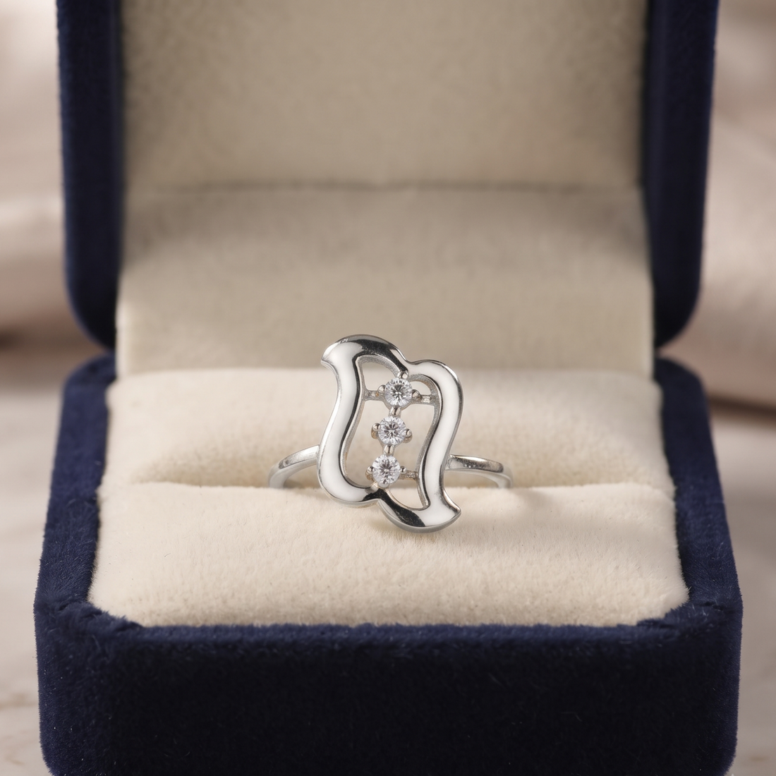 A silver ring with three small clear stones set in an abstract open-loop design, displayed inside an open navy blue velvet jewelry box with a cream-colored cushion.
