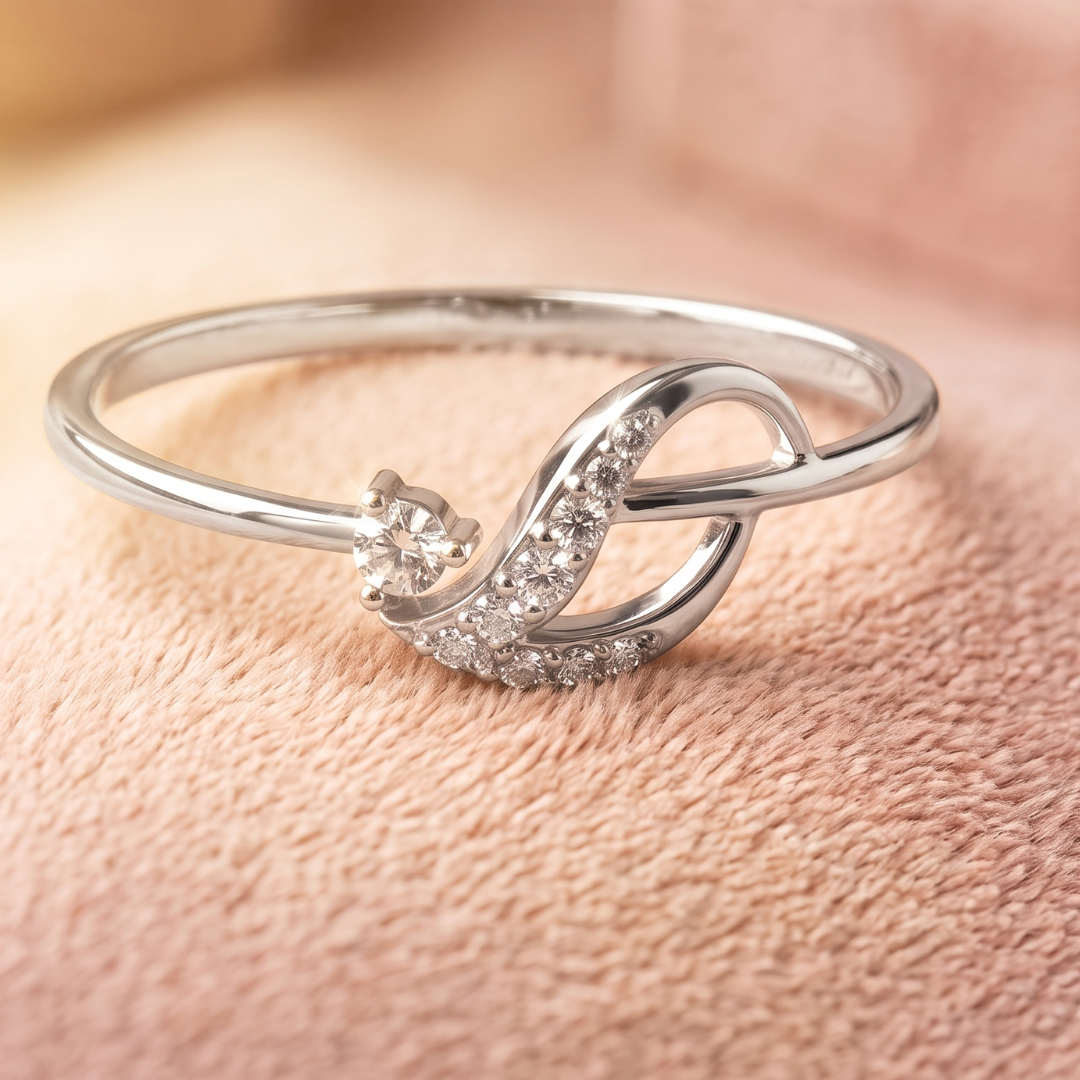 A silver open band ring with a curved infinity-style design, accented by small clear stones and a central clear gemstone, photographed on a soft pink textured surface.