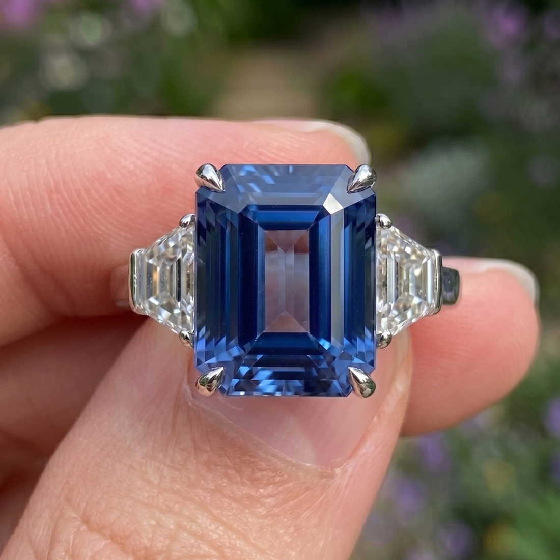 A close-up of a ring being held between fingers, featuring a large emerald-cut blue sapphire center stone with baguette-cut clear side stones in a silver-toned setting, against a blurred green and purple outdoor background.
