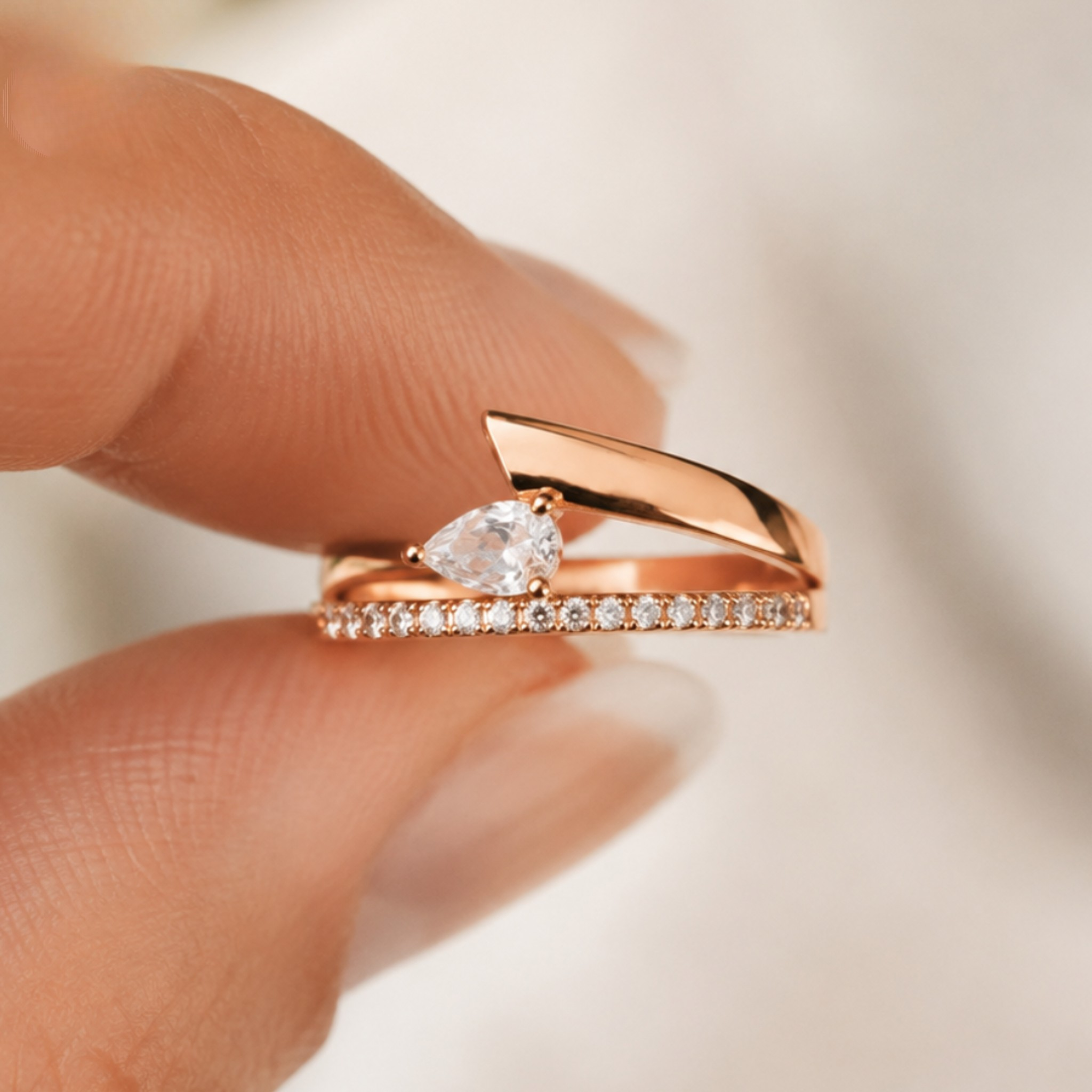 A close-up of a hand holding a rose gold ring set with a pear-cut clear gemstone and a pavé band, against a soft, out-of-focus light background.