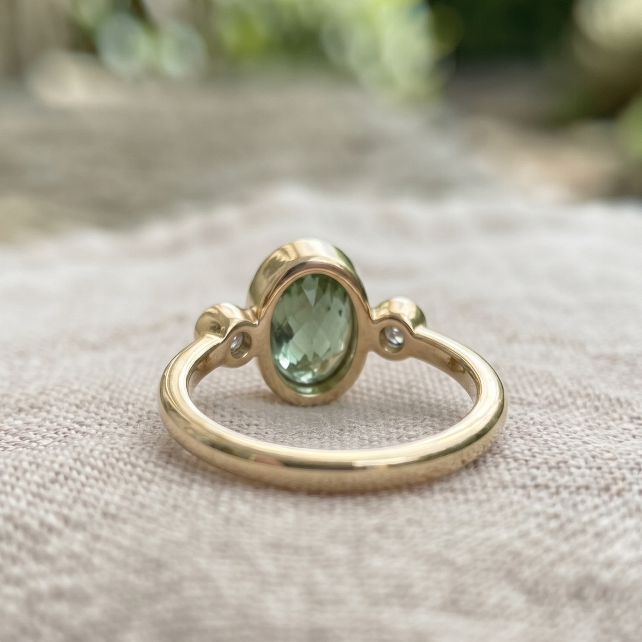 A gold ring with an oval green gemstone and a small clear accent stone, resting on a textured beige fabric surface with a blurred outdoor background.