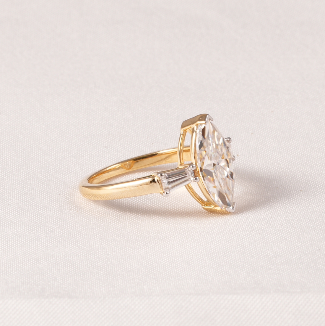 A yellow gold ring featuring a large marquise-cut clear gemstone in a prong setting, accented by a smaller round diamond on the band, set against a textured off-white background.
