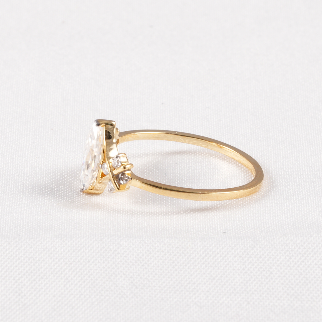 A yellow gold ring with a marquise-cut clear gemstone and a small accent stone, shown on a textured white background.