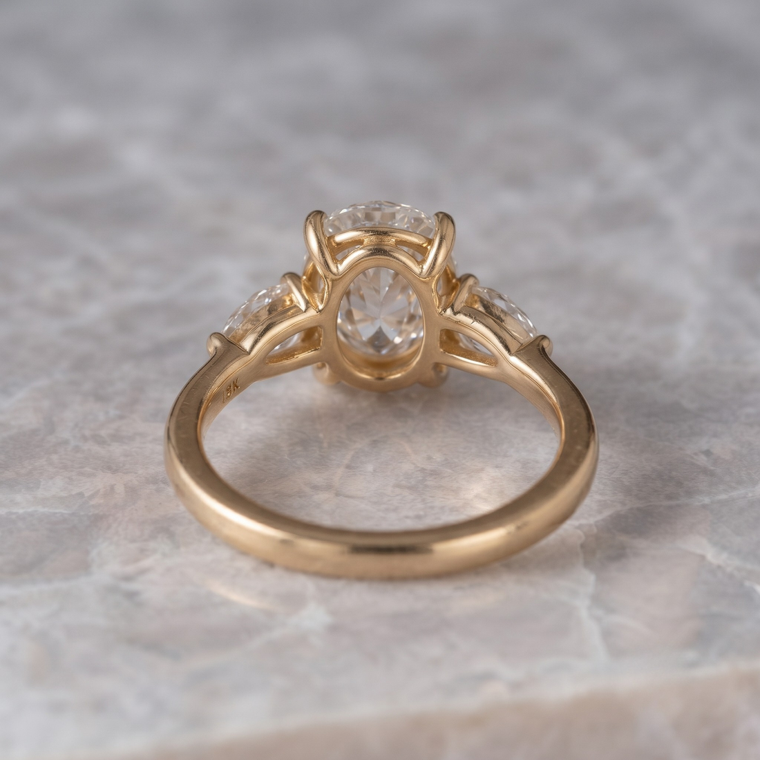 A yellow gold ring with a central round-cut clear gemstone and two smaller side stones, photographed on a light stone-textured surface.