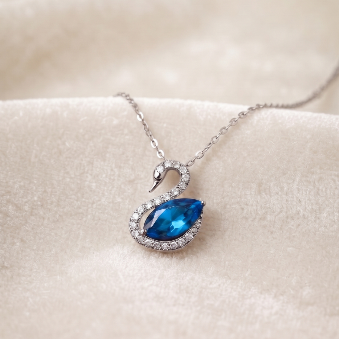 A silver-tone swan pendant necklace featuring a faceted blue gemstone and clear crystal accents, resting on a soft beige fabric surface.