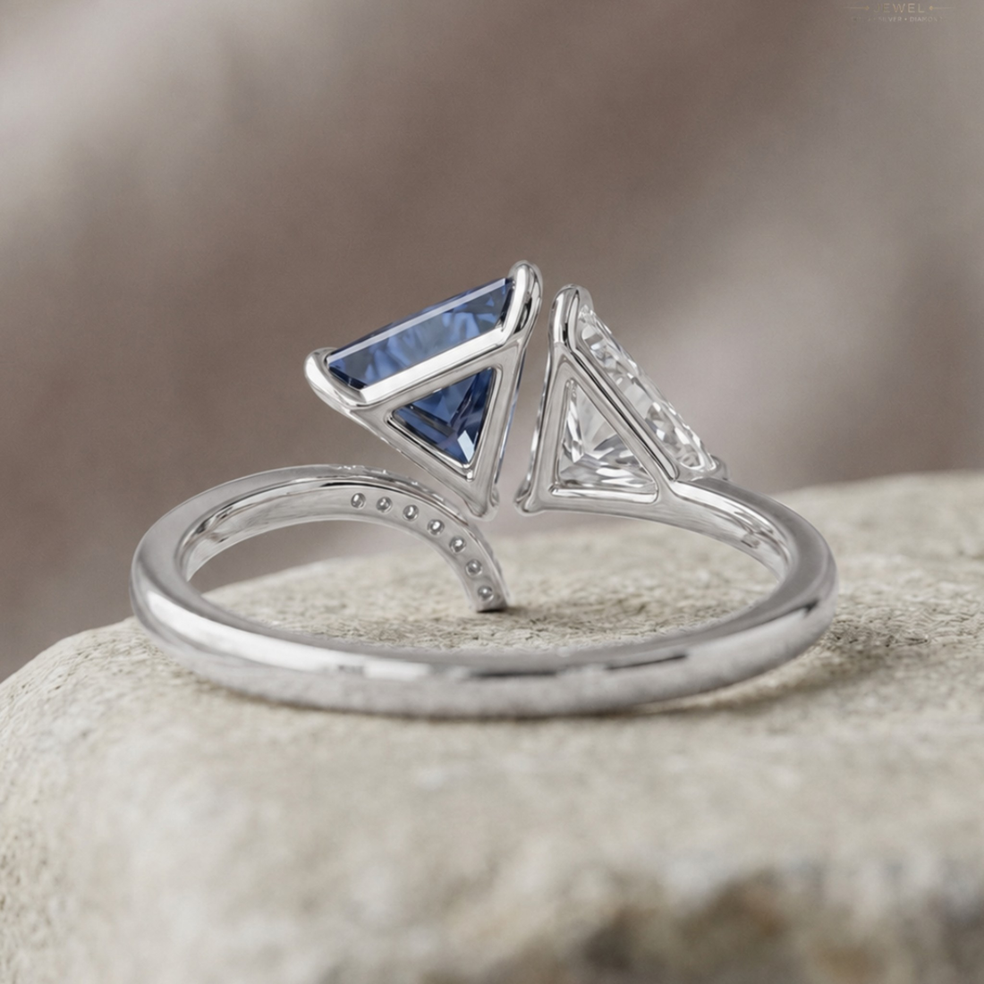 A silver-toned triangular split-shank ring with a blue princess-cut center stone and a small clear gemstone, resting on a textured stone surface.