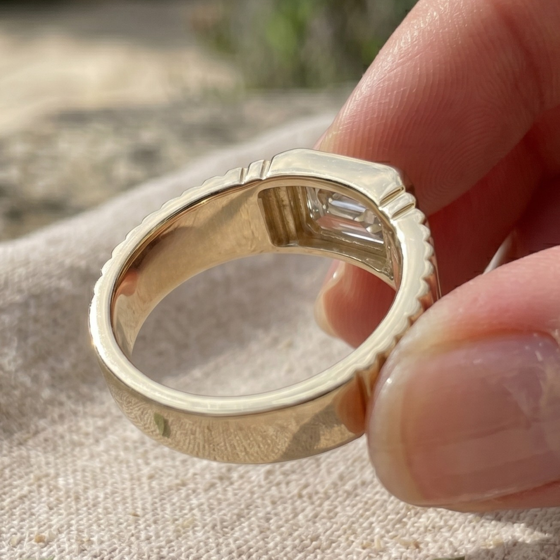 A close-up of a gold ring with a rectangular clear gemstone, held between fingers against a soft beige fabric background.