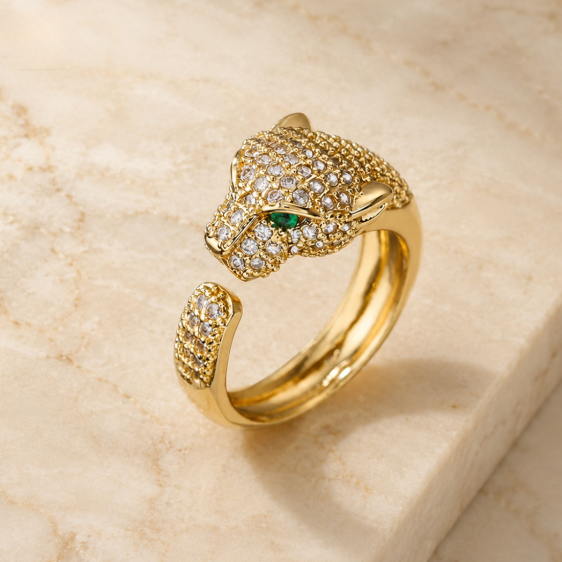 Gold-toned open ring shaped like a panther head, set with pavé clear stones and a small green eye accent, photographed on a light beige stone surface.