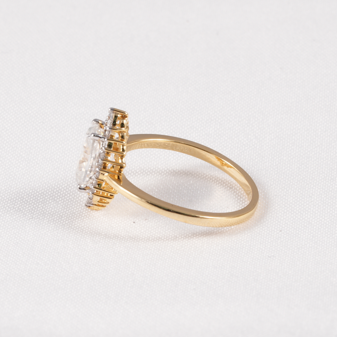 A yellow gold ring with a large clear round-cut center stone and smaller clear stones along the halo, photographed on a white textured background.