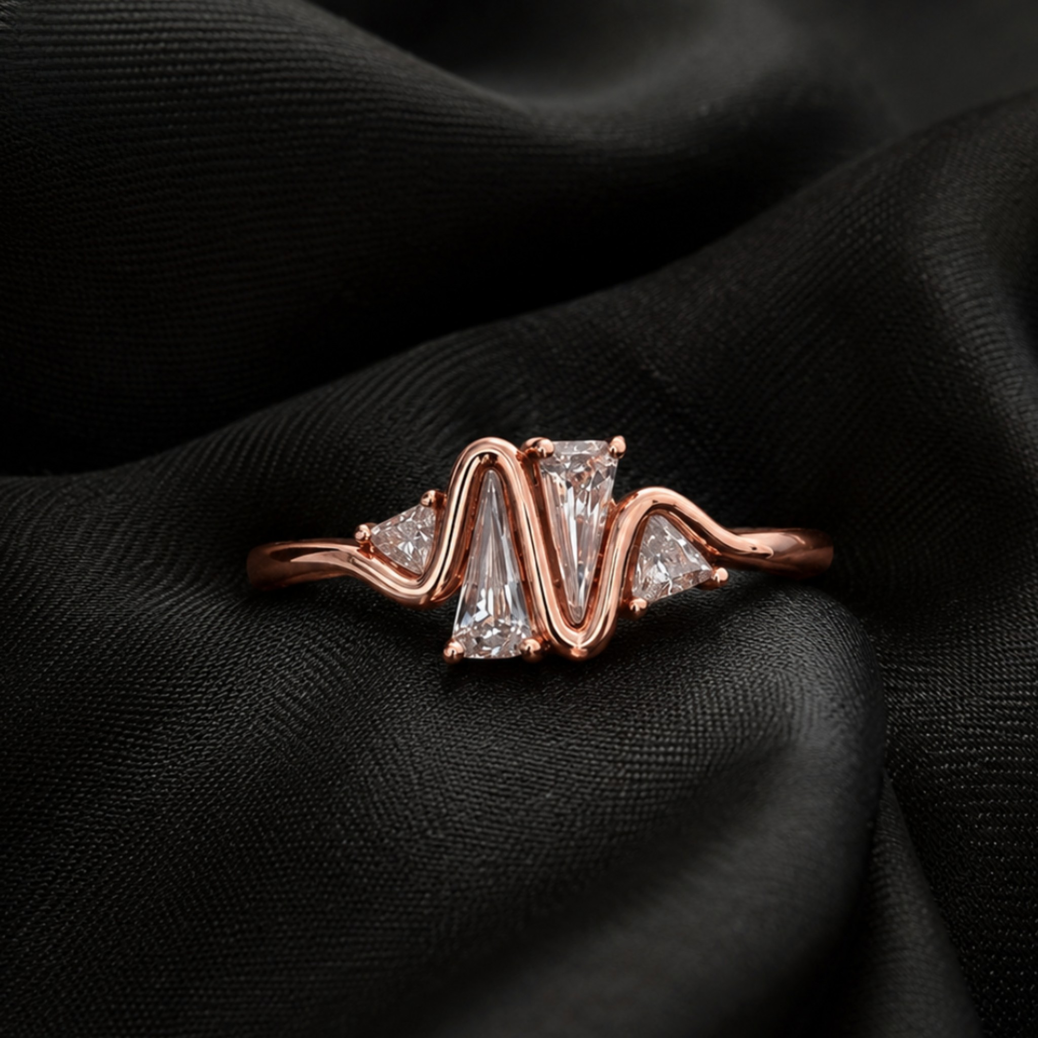 A rose gold ring with a wavy, zigzag design featuring a central rectangular clear gemstone and smaller side stones, displayed on a draped black fabric background.