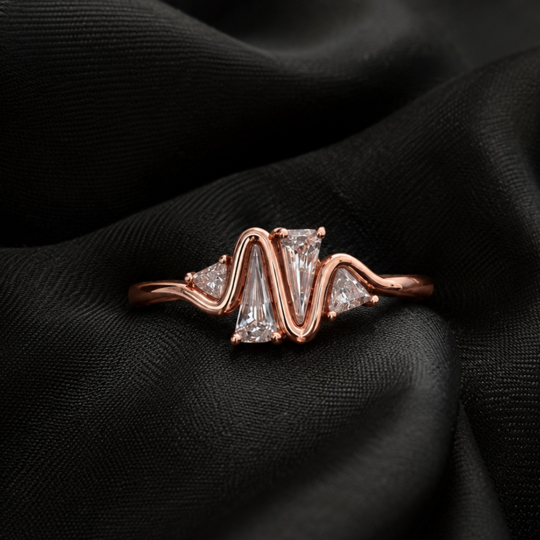 A rose gold ring with a wavy, zigzag design featuring a central rectangular clear gemstone and smaller side stones, displayed on a draped black fabric background.