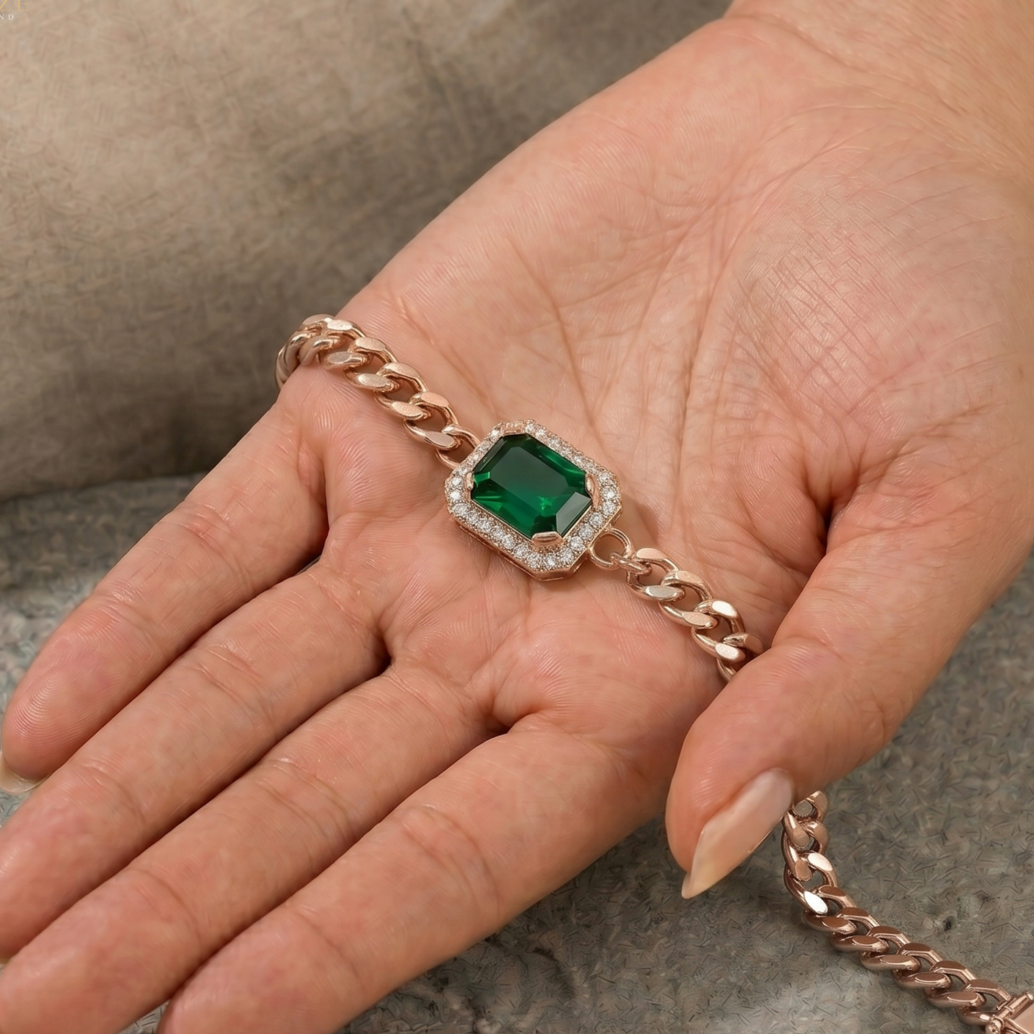 A close-up of a hand holding a rose gold chain bracelet with a square-cut green emerald center stone surrounded by a halo of small clear stones.