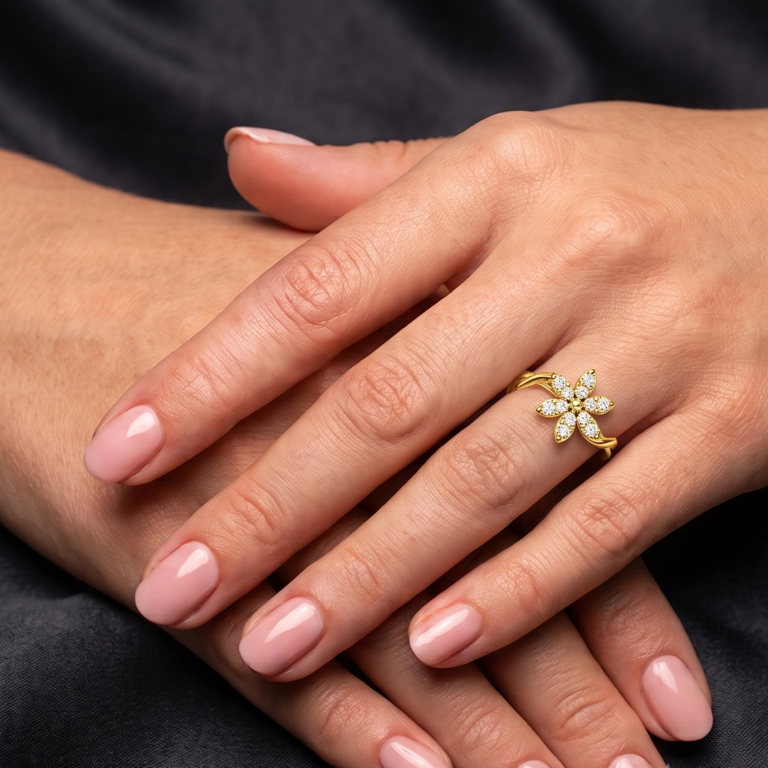 A close-up of a person's hands with light pink manicured nails, wearing a gold ring shaped like a small flower with clear stones, set against a dark fabric background.