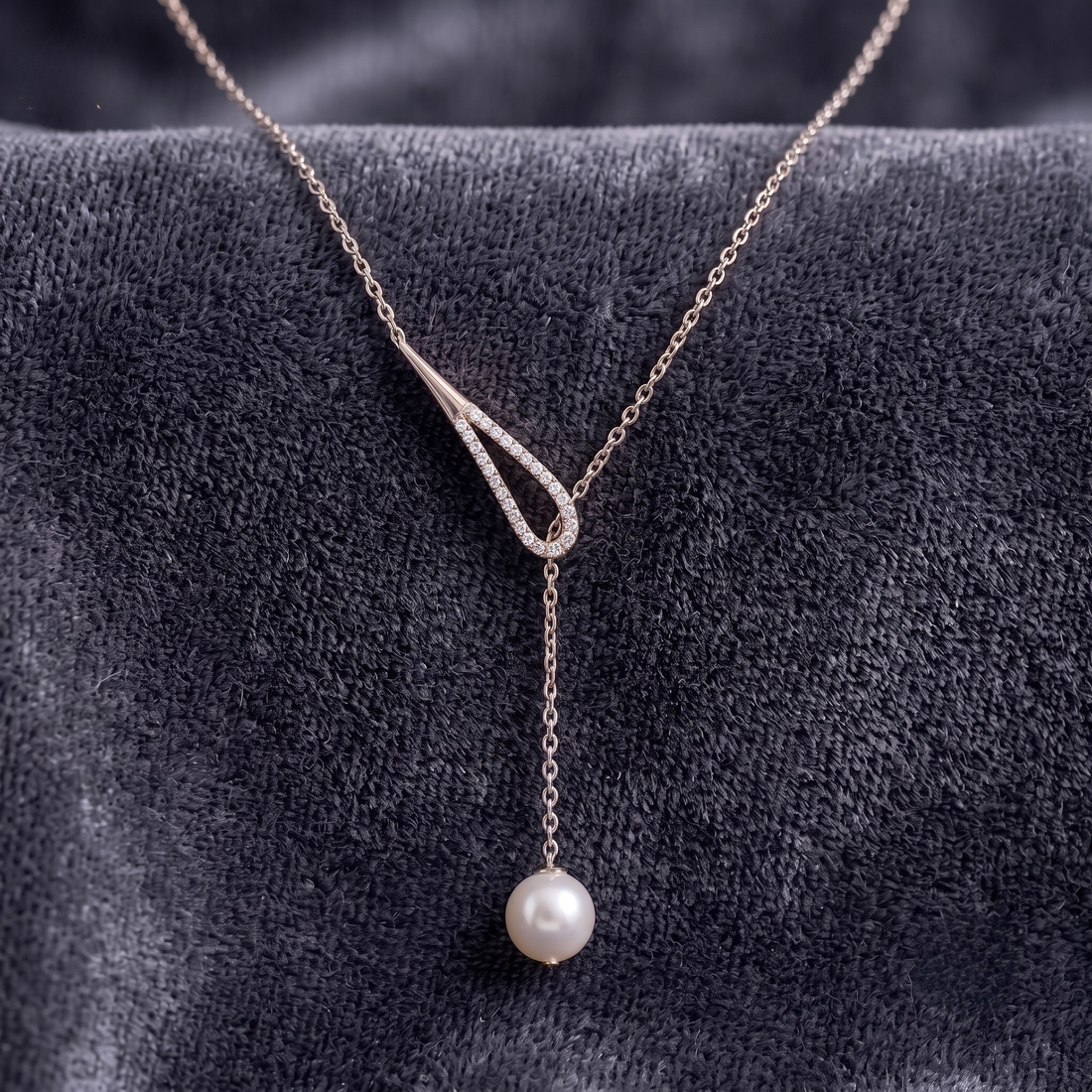 A delicate Y-shaped necklace with a white pearl pendant, displayed on a textured dark gray velvet surface. The necklace features a fine silver-toned chain with a split V-design, where one side is a simple loop and the other is adorned with small clear stones, culminating in a single drop chain ending in a round white pearl.