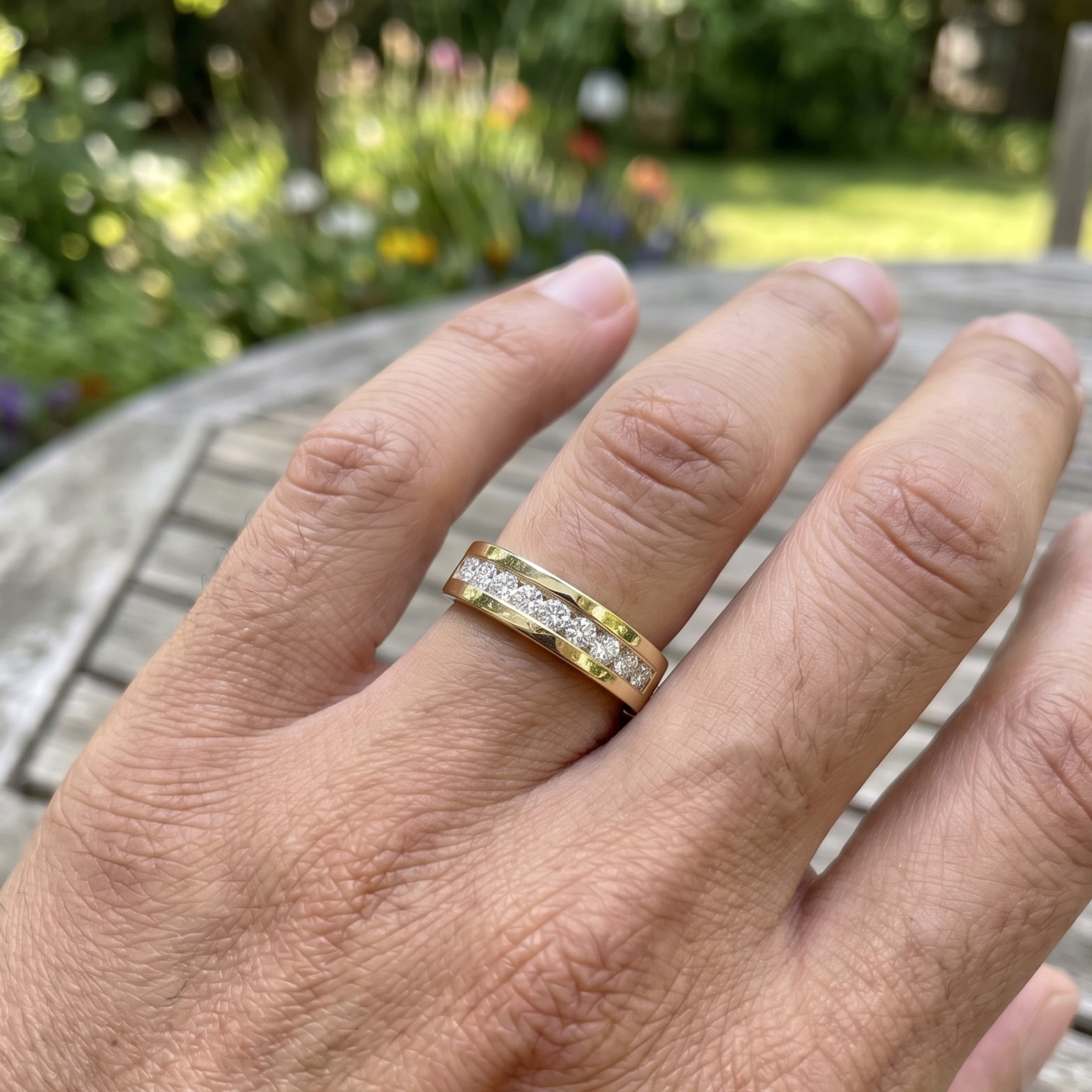 A yellow gold band ring with a row of small clear stones, shown worn on a person's hand in an outdoor garden setting.