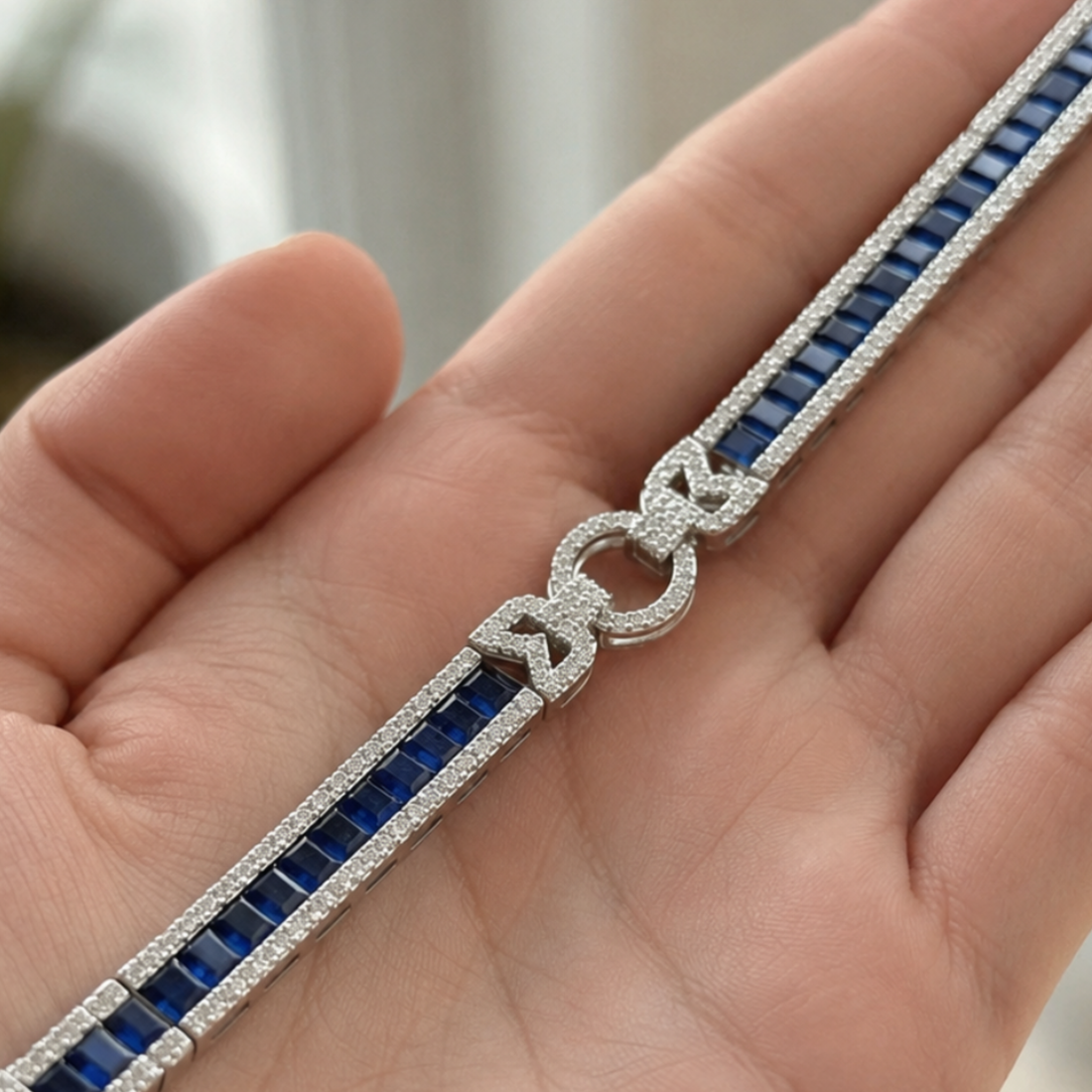 A close-up of a silver-toned bracelet with deep blue rectangular stones and pavé-set clear stones, shown resting on a person's hand.