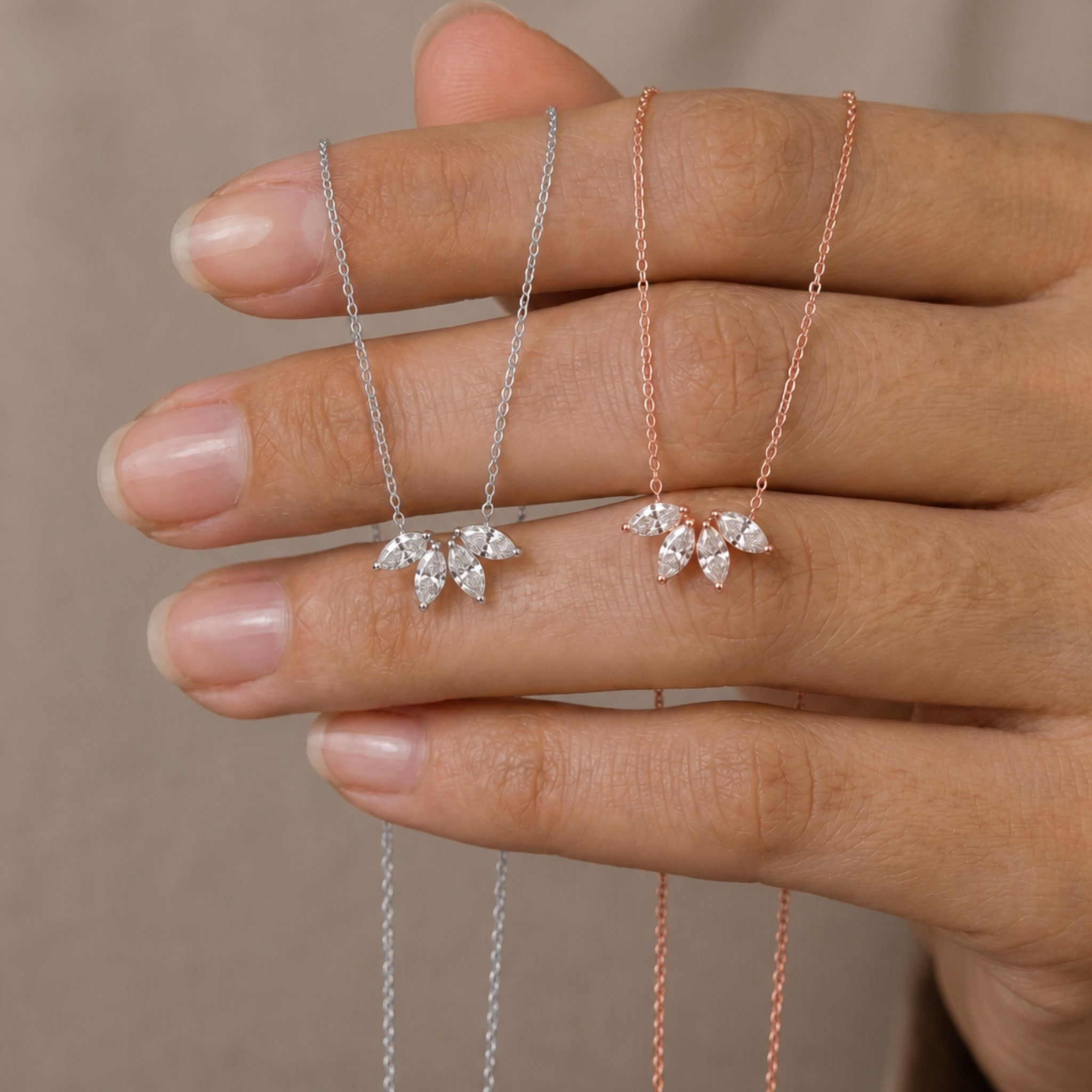 Two delicate leaf-shaped necklaces with clear crystal-like stones, one in silver and one in rose gold, displayed on a person's hand against a soft beige background.