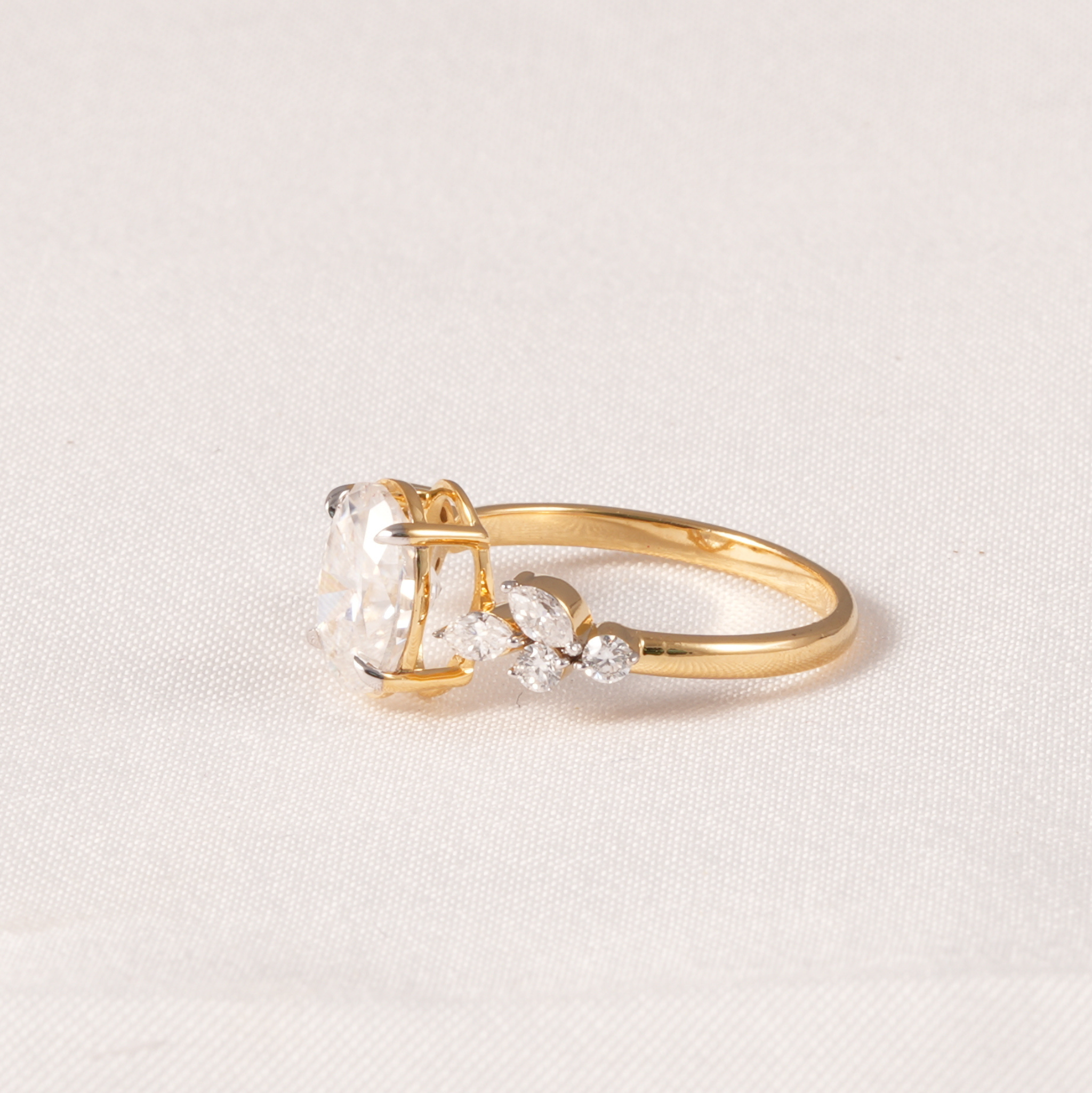 A yellow gold ring featuring a large clear rectangular gemstone in a prong setting, accented by small marquise and round clear stones on the band, set against a light beige textured fabric background.