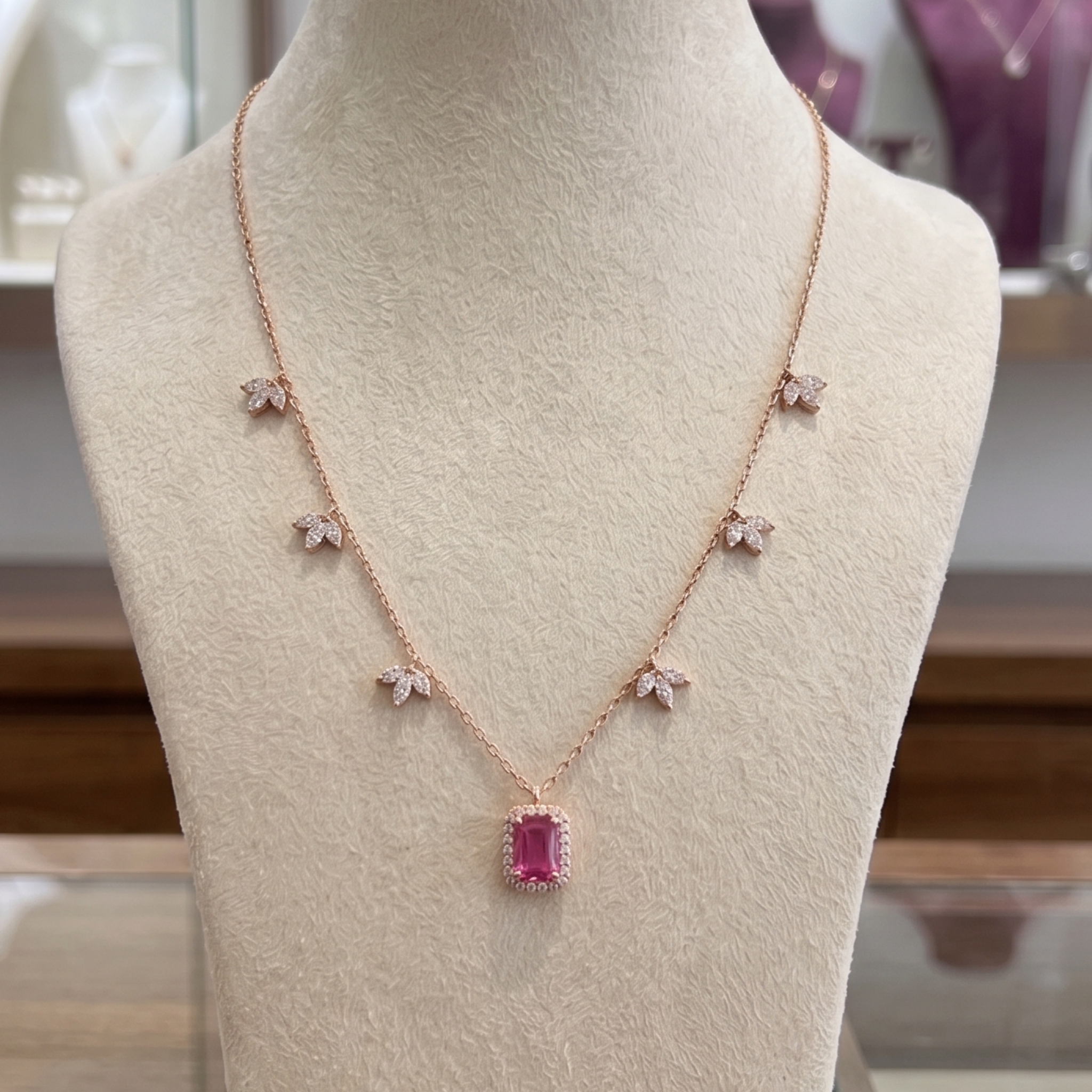 A rose gold necklace displayed on a beige jewelry bust. The necklace features a central rectangular pink gemstone pendant surrounded by small clear stones, with five matching small flower motifs spaced along the chain.