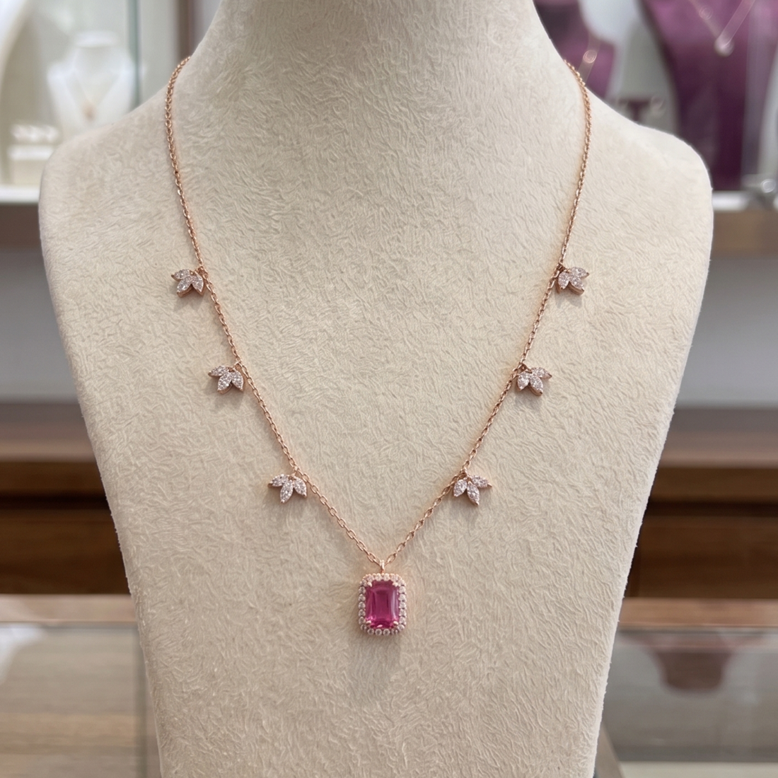 A rose gold necklace displayed on a beige jewelry bust. The necklace features a central rectangular pink gemstone pendant surrounded by small clear stones, with five matching small flower motifs spaced along the chain.