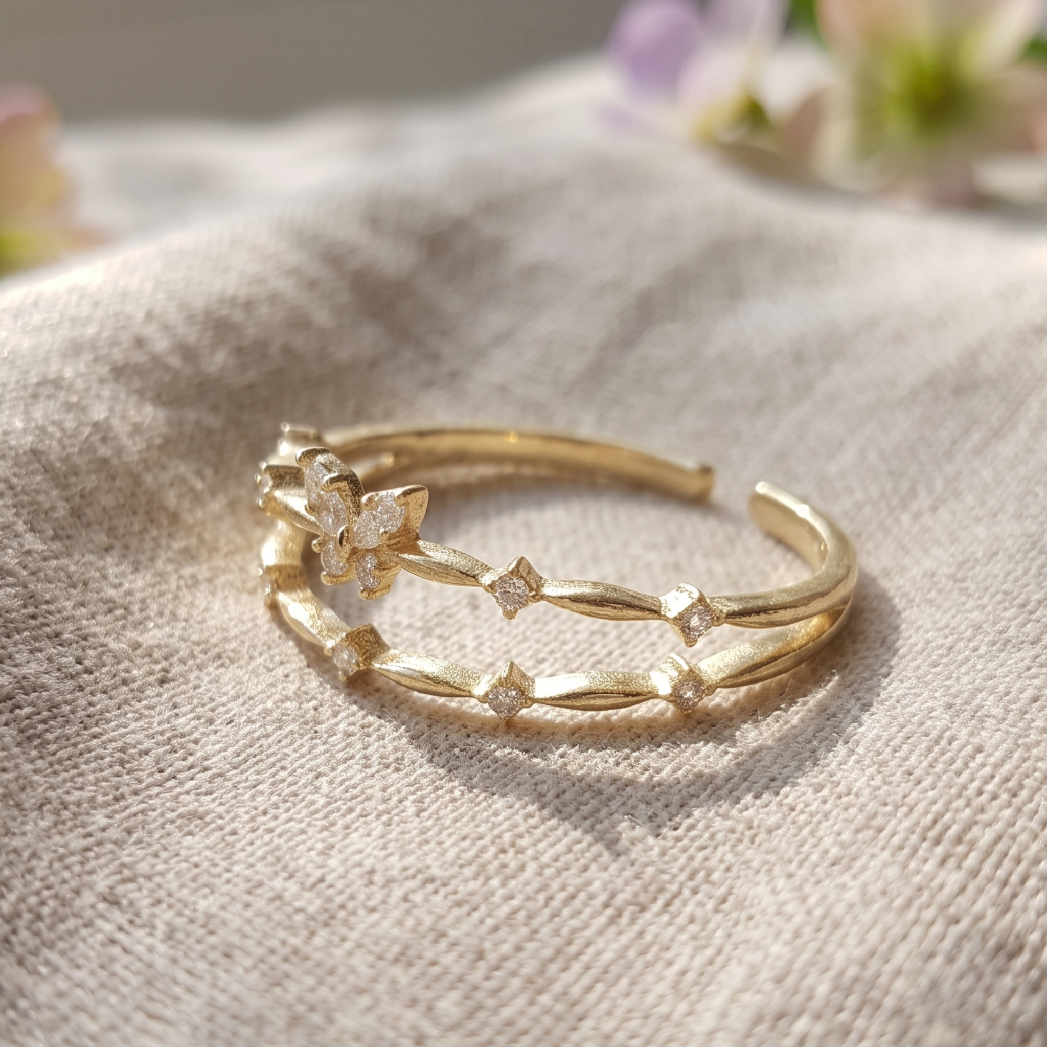 A delicate gold open bangle ring featuring a small flower motif and small clear crystal accents, resting on a textured beige linen fabric with a soft-focus background.
