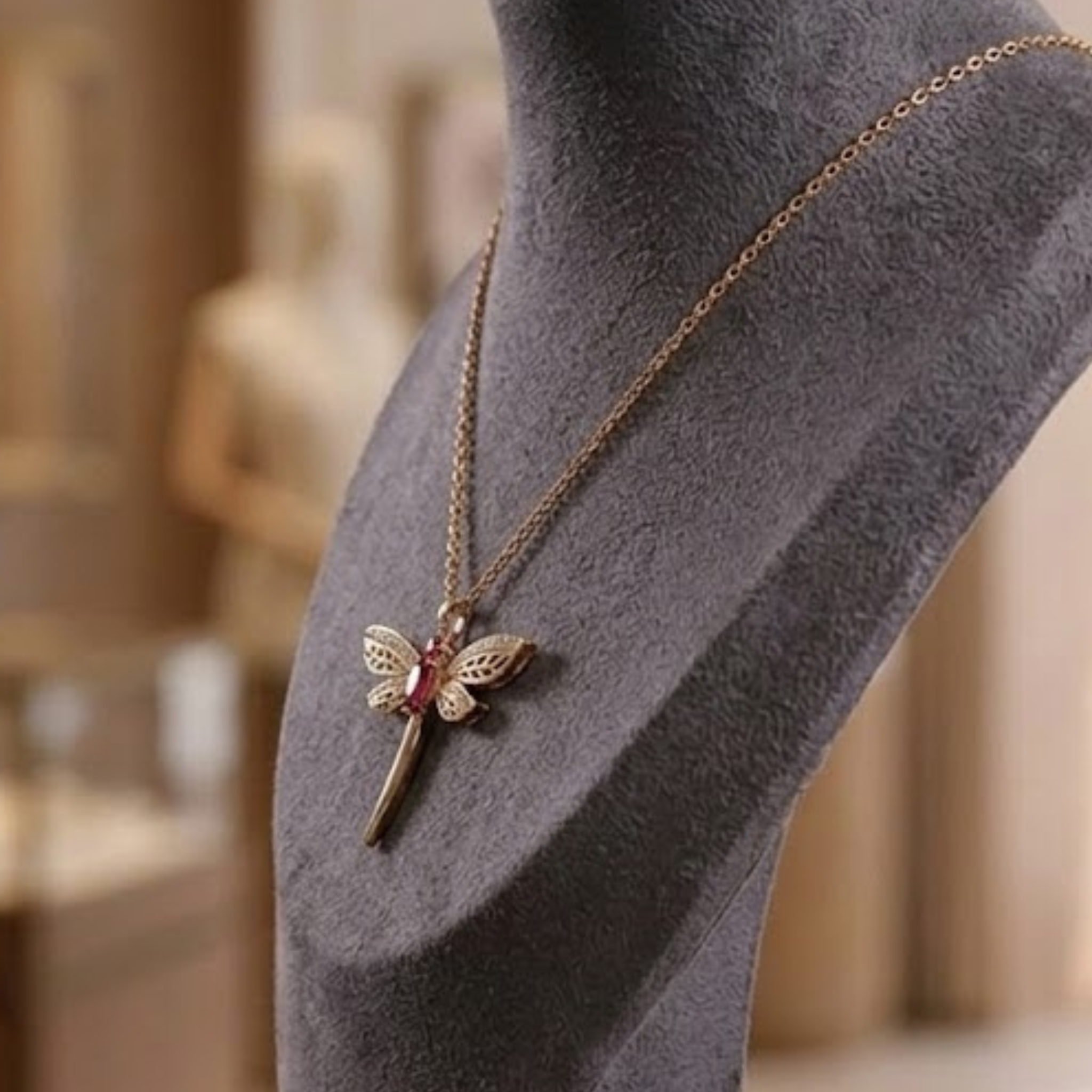 A gold dragonfly pendant necklace with a small red gemstone accent, displayed on a gray velvet jewelry bust.