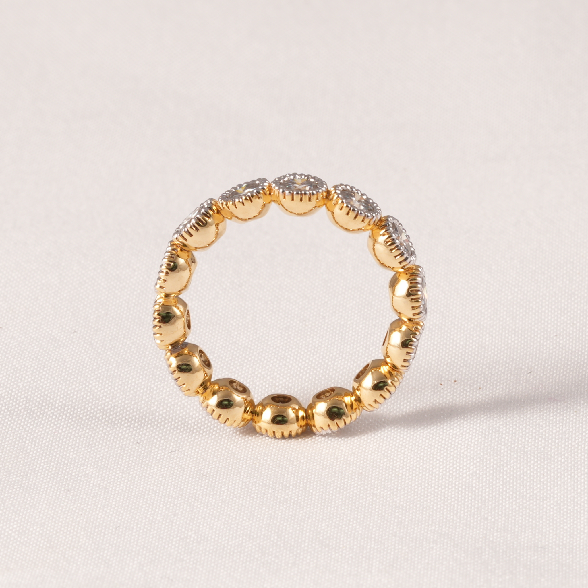 A gold beaded ring with alternating smooth round beads and textured dome beads set with small clear stones, photographed on a light beige fabric background.