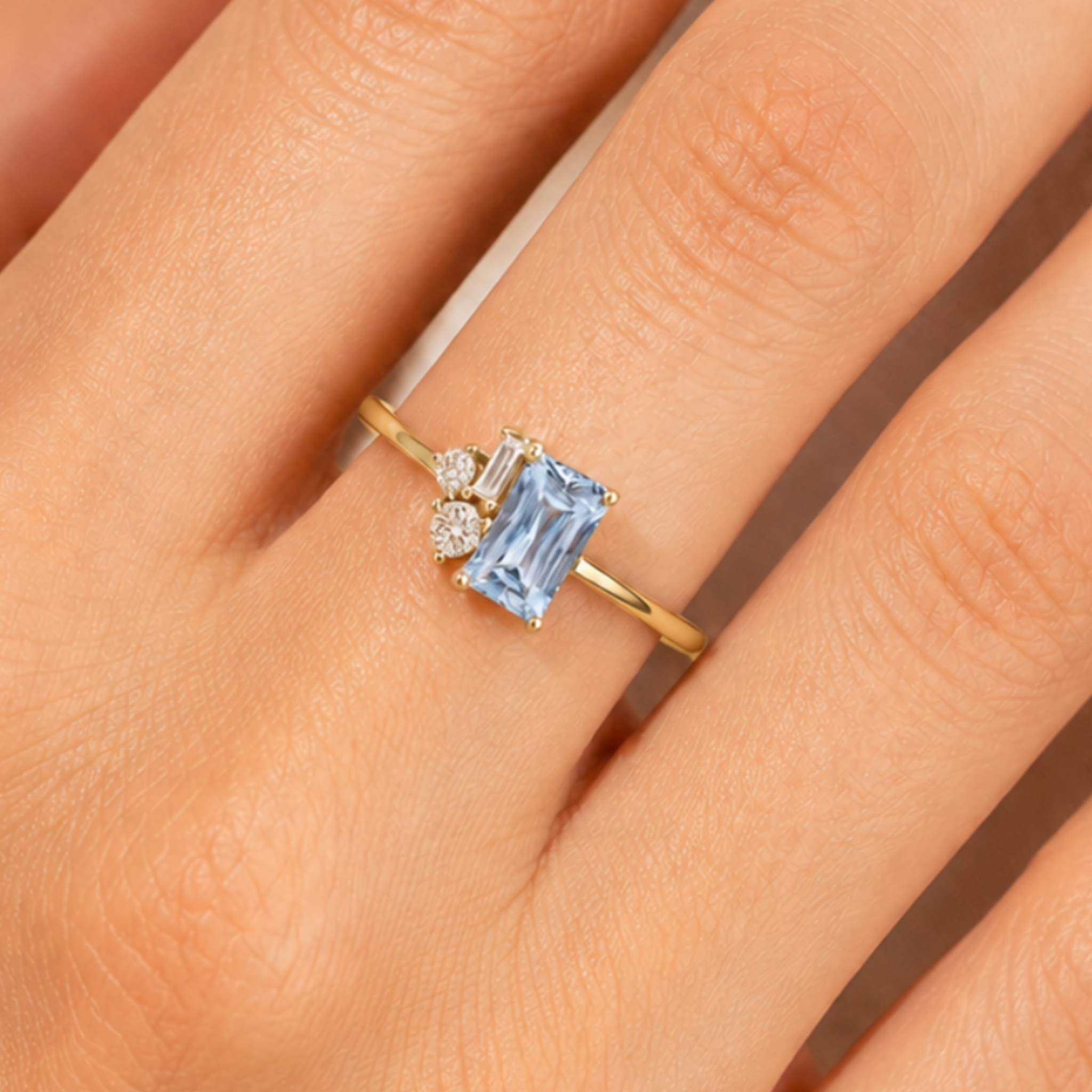A gold ring with a rectangular light blue gemstone and two small clear accent stones, shown worn on a person's finger.