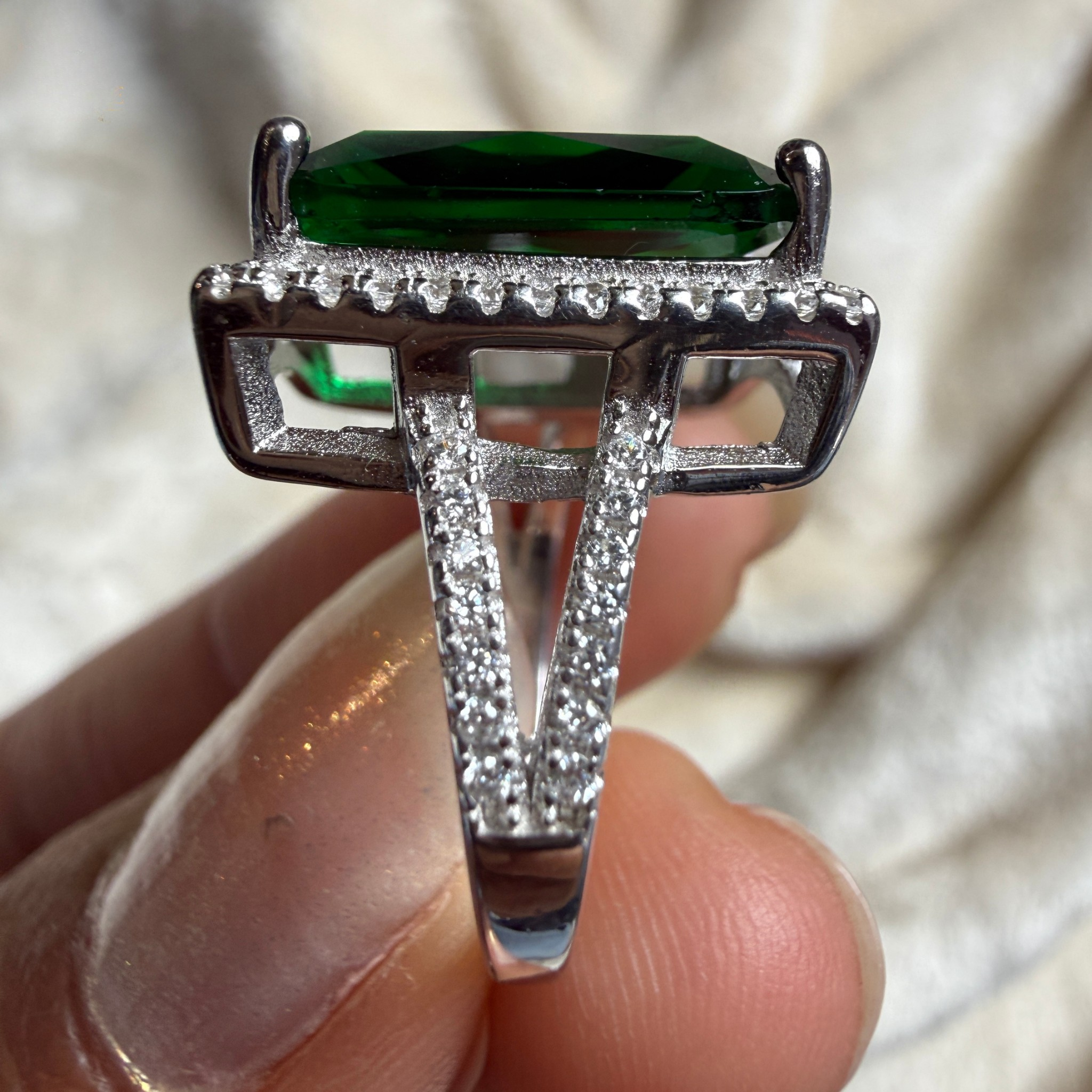 A close-up of a silver ring held between fingers, featuring a rectangular green gemstone in a prong setting with pavé diamonds along the band and shoulders.