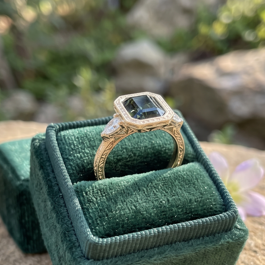 A gold ring with a large dark green rectangular gemstone and smaller side stones, displayed in an open dark green velvet ring box outdoors with a blurred natural background.