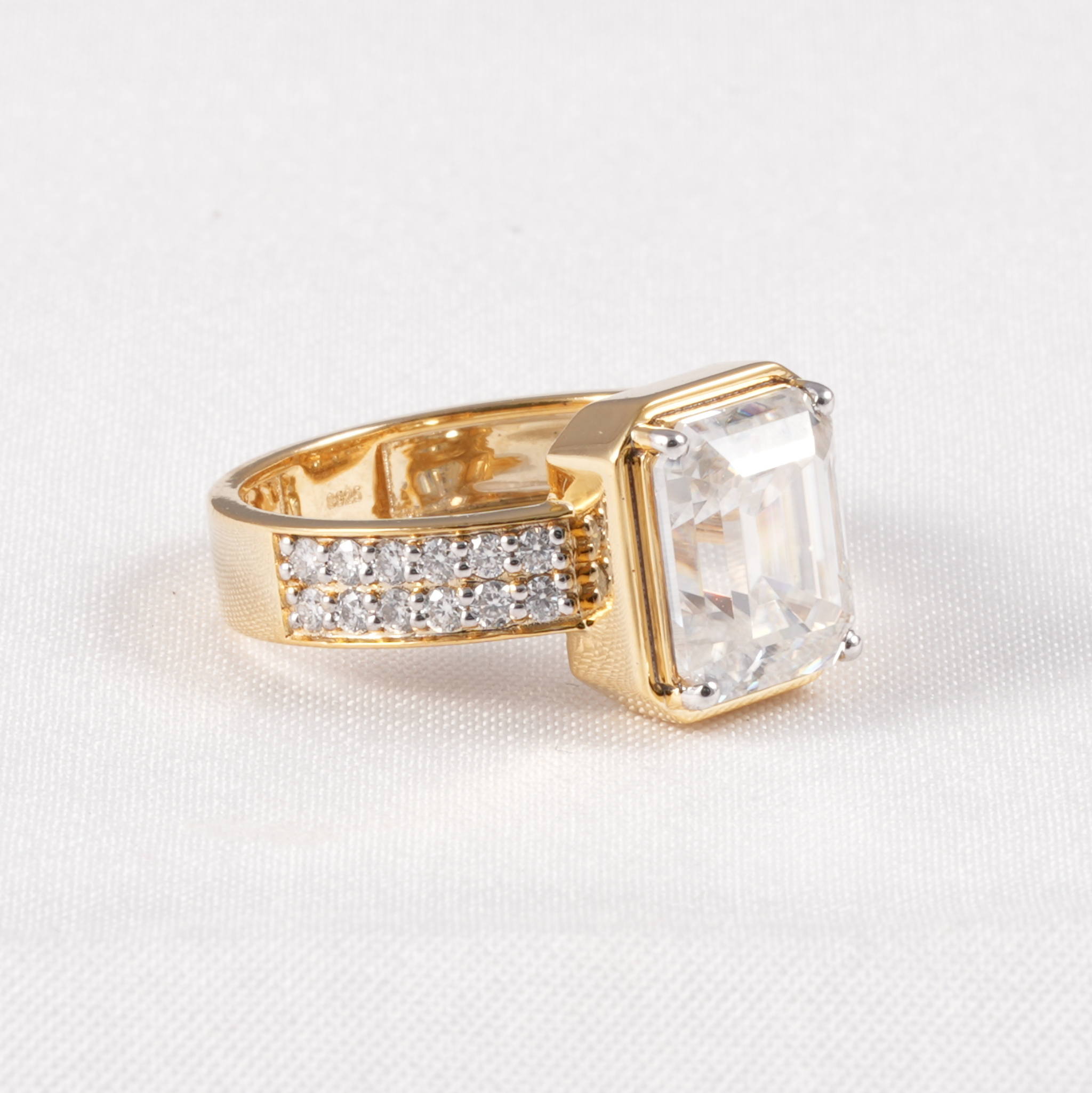 A yellow gold ring featuring a large rectangular clear gemstone and a pavé band with small clear stones, set against a white textured background.