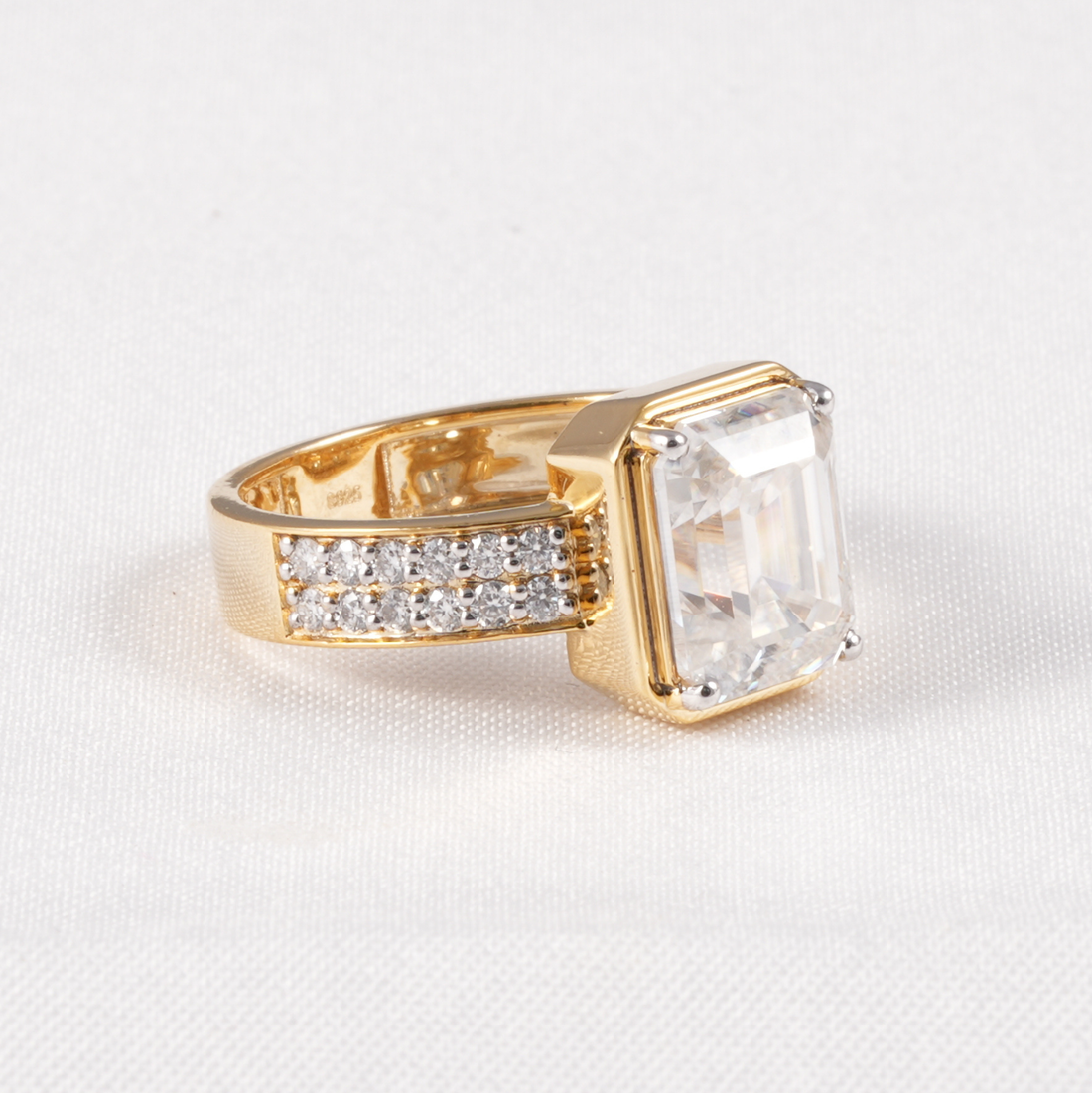 A yellow gold ring featuring a large rectangular clear gemstone and a pavé band with small clear stones, set against a white textured background.