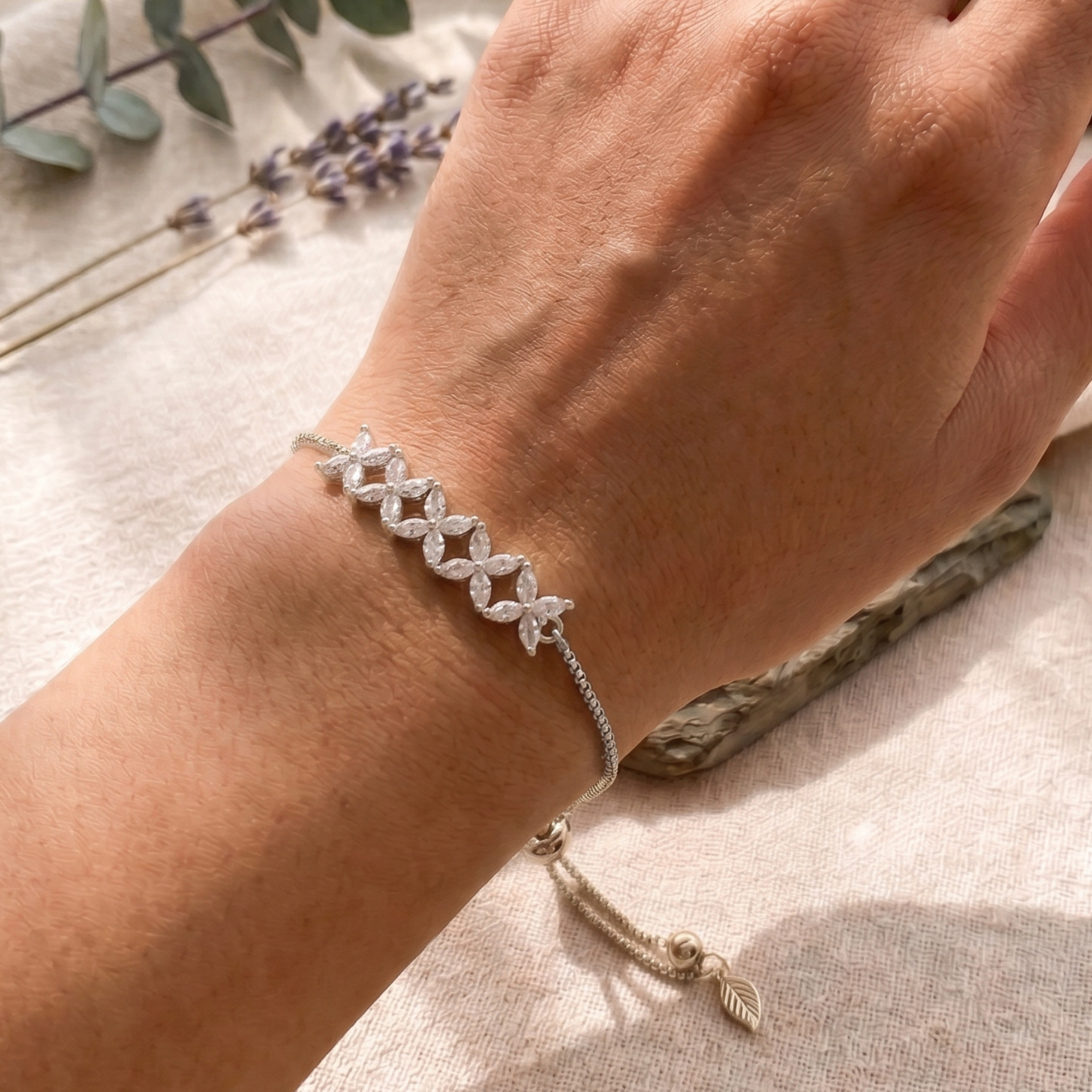 A delicate silver chain bracelet with four interlocking flower motifs and a small tassel charm, shown worn on a person's wrist against a soft neutral fabric background.