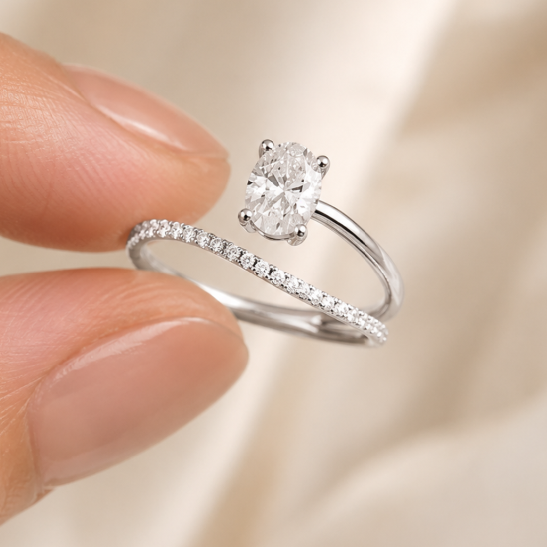 A silver solitaire engagement ring with a round-cut clear center stone and a pavé-set band, shown being held between two fingers against a soft beige fabric background.