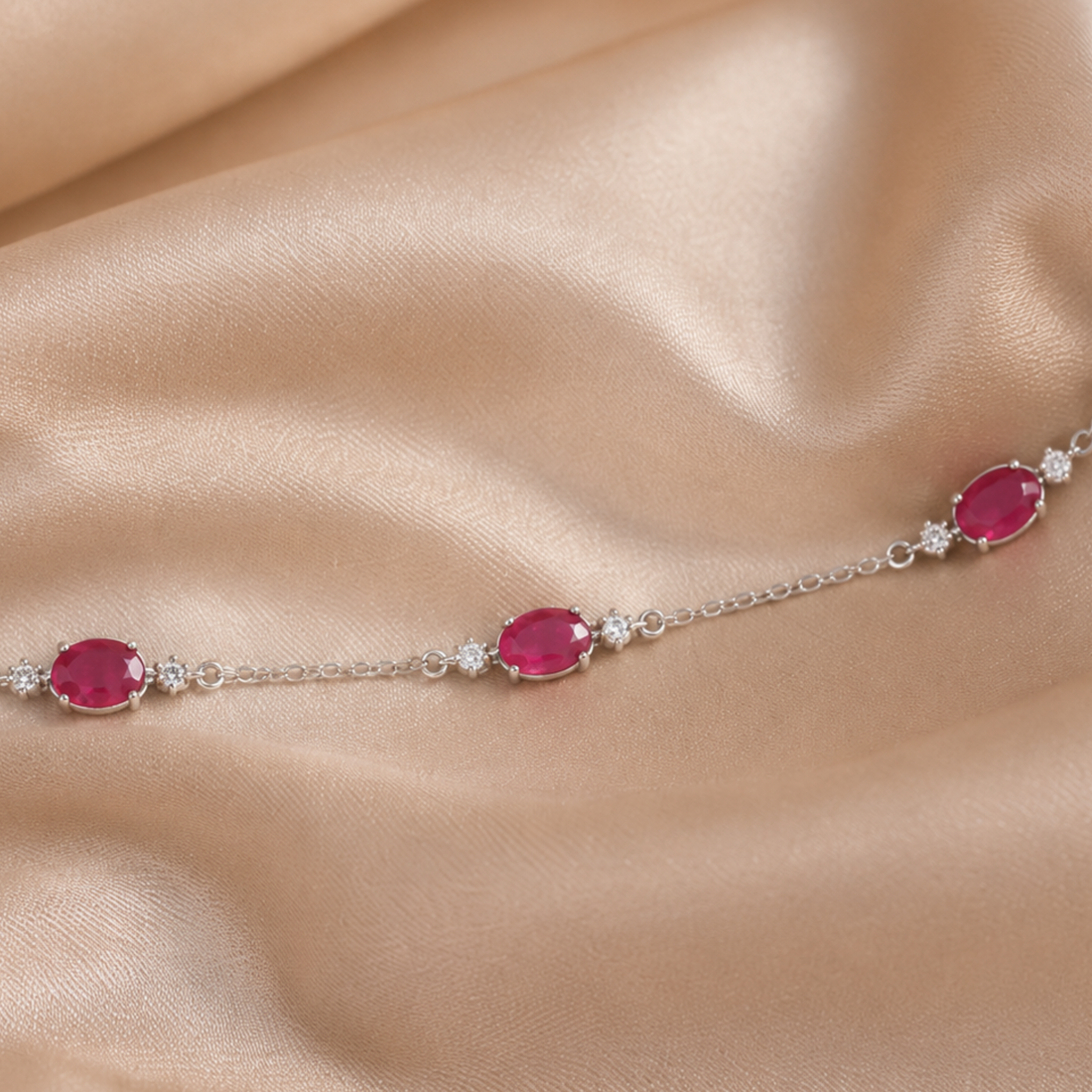 A delicate silver chain bracelet featuring three oval pinkish-red gemstones and small clear stones, displayed on a draped beige satin fabric.