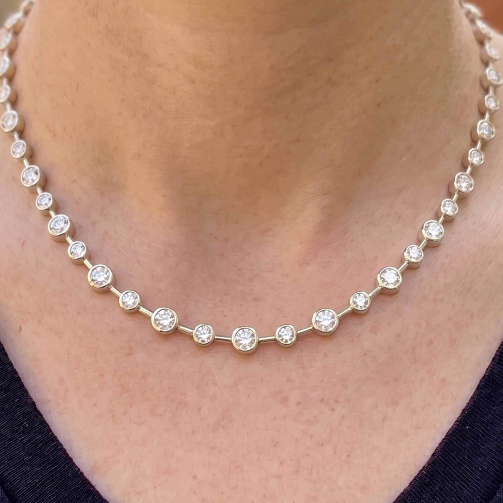A close-up of a delicate gold tennis necklace with clear round-cut gemstones worn on a person's neck.