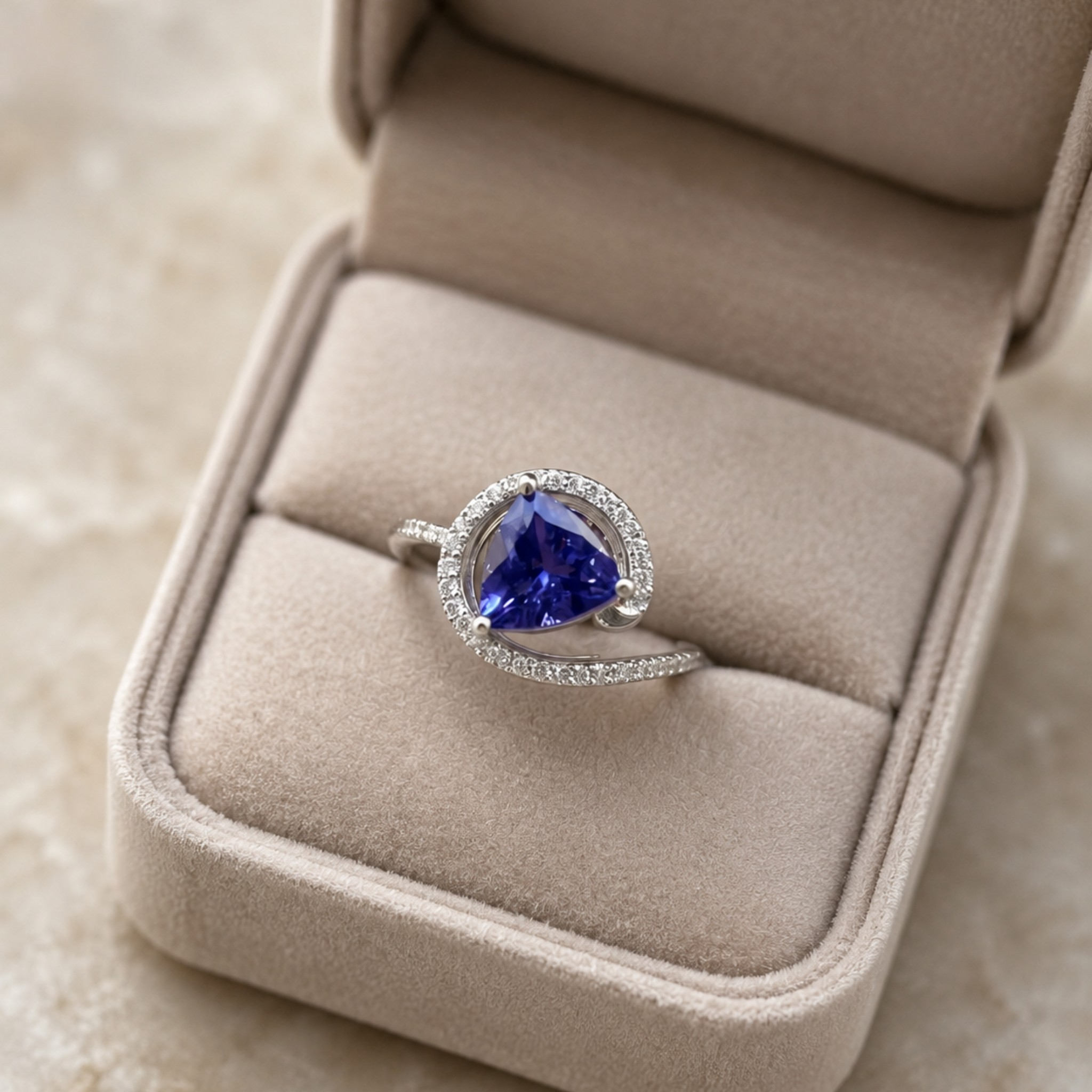 A heart-shaped blue gemstone ring with a halo of small clear stones and a curved silver band, displayed in an open beige jewelry box.